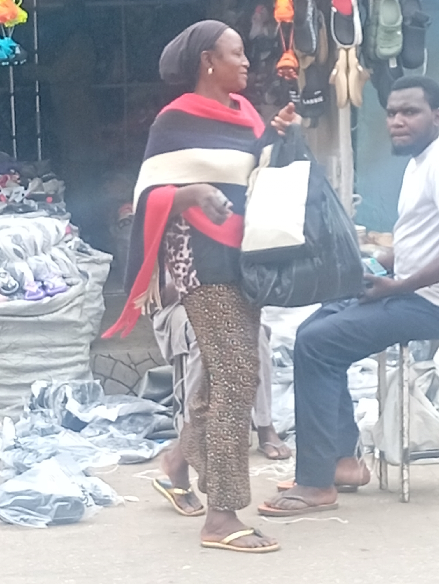 In Maiburiji, Nigeria, an outdoor scene depicts a woman with a head covering, draped in a red, white, and black shawl. She holds a large black and white bag. She is wearing patterned trousers and yellow sandals. Behind her, a man is seated, wearing a white shirt and blue trousers, using a mobile phone. In the background, there is merchandise including sandals hanging and piles of packaged items.