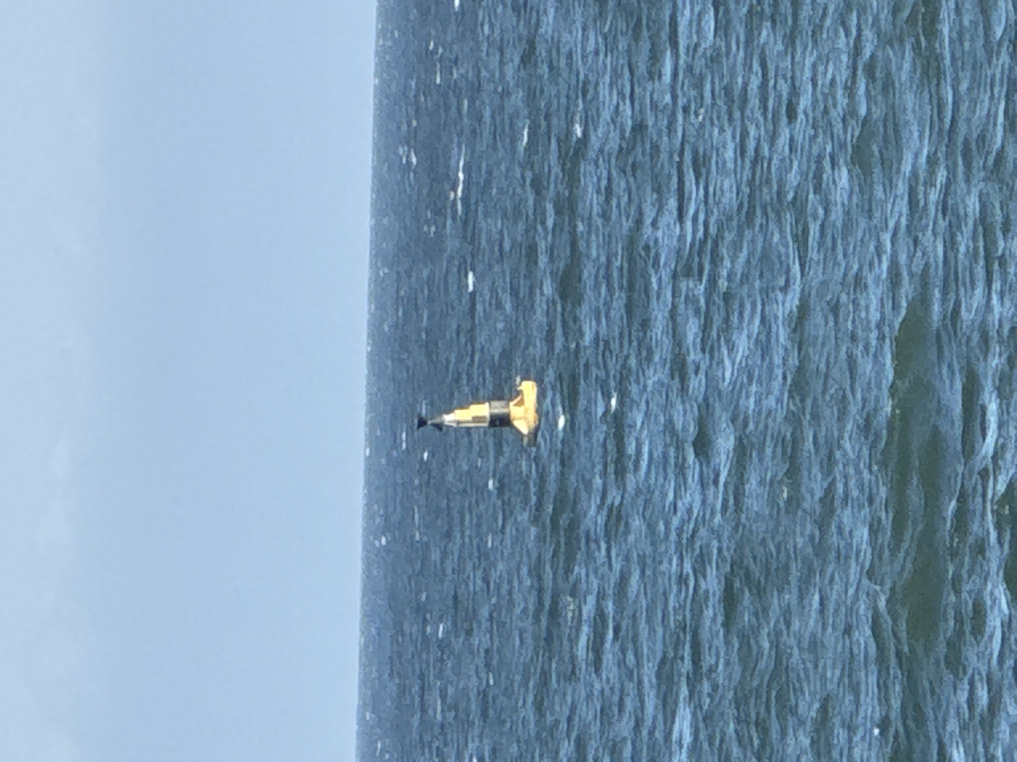 A yellow and black buoy floats on the surface of the choppy, dark blue ocean under a hazy sky. The buoy has a conical top with a black tip and a yellow body, banded with black near the top. The horizon line, where the sea meets the sky, is visible in the background. The image appears to be taken from a distance, possibly from a shore or a boat. The location is not specified in Niterói, Brazil.