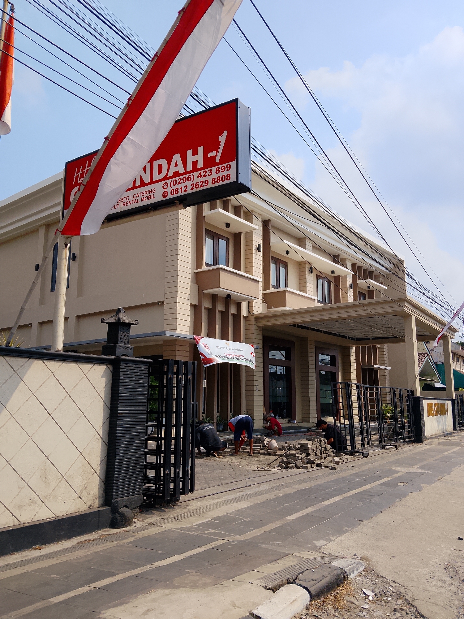 The image depicts the exterior of a multi-story building with a beige and brown facade, identified by a red sign as "HOTEL SUNDA-1". The hotel offers "RESTAURANT | CATERING | PUTRI RENTAL MOBIL". Two phone numbers are listed on the sign: (0296) 423 899 and 0812 2629 8808. The building has several windows and balconies. In front of the building, there is a black metal fence and a white tiled wall. Several people are working on the pavement in front of the gate, laying down bricks. A red and white Indonesian flag is draped over electrical wires above the building. The scene appears to be located on a street in the city of Balun, Indonesia, based on the text on the building's sign. The sky is clear with a few scattered clouds. The overall impression is of a commercial establishment undergoing some form of maintenance or construction.
