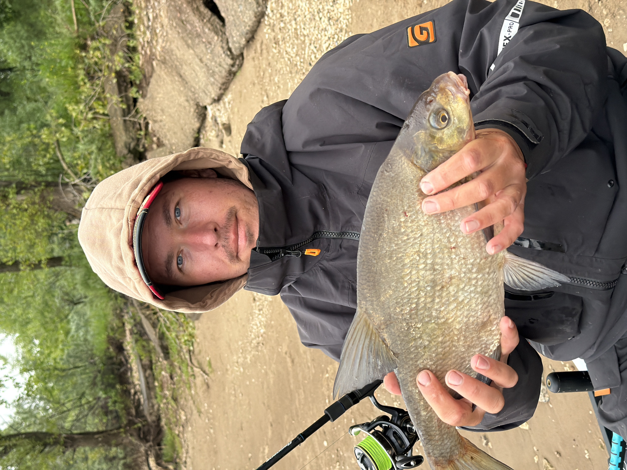 The image, rotated 90 degrees clockwise, captures a man holding a large fish against an outdoor backdrop. The man, an adult with striking blue eyes, looks directly at the viewer with a neutral to slightly pleased expression. He is wearing a dark grey or black hooded jacket with the light brown hood pulled over his head, revealing a black cap with a red brim underneath.
He presents a large, silvery-brown fish, held horizontally with both hands. The fish appears to be a freshwater species, possibly a bream or carp, characterized by its prominent scales and shape. Below the fish, in the man's left hand, a black fishing rod with a reel spooled with bright lime-green line is visible, confirming a fishing activity.
The setting is a natural, somewhat rugged environment, likely a riverbank or lake shore. The ground beneath him is sandy and rocky, a light brown color. In the background to the left, lush green foliage from trees or bushes can be seen. To the right, there are large, textured formations that could be concrete blocks or natural rocks.
Visible text and logos include an orange, stylized "G" logo on the man's left chest area of his jacket. On his right sleeve, the words "FLEX-PRO" are printed alongside a white barcode-like design. The cool weather suggested by the jacket implies it's either an overcast day, early morning, or late afternoon in Noviy, Russia.