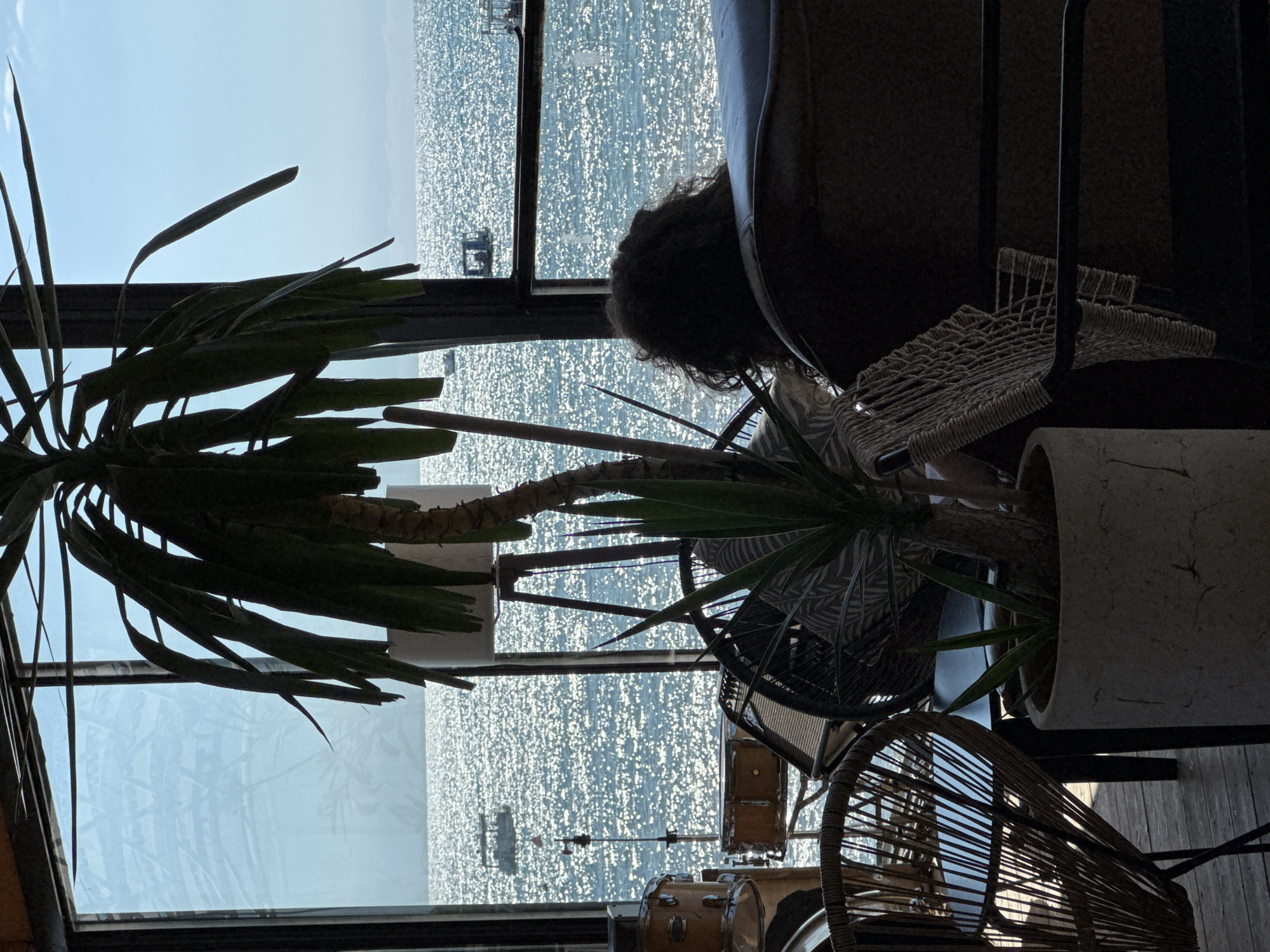 The image shows an indoor scene with a large window overlooking the sea. The window is partially obscured by a potted plant and several pieces of furniture. The sea is visible in the background, with sunlight glinting off the water and several boats in the distance. The room appears to be a cafe or restaurant, with wicker chairs and a table visible. A person with dark, curly hair is seated in a chair, facing away from the camera and towards the sea. Drums and an amplifier are also visible in the foreground, suggesting the space might also be used for musical performances. The location is identified as Sochi, Russia.