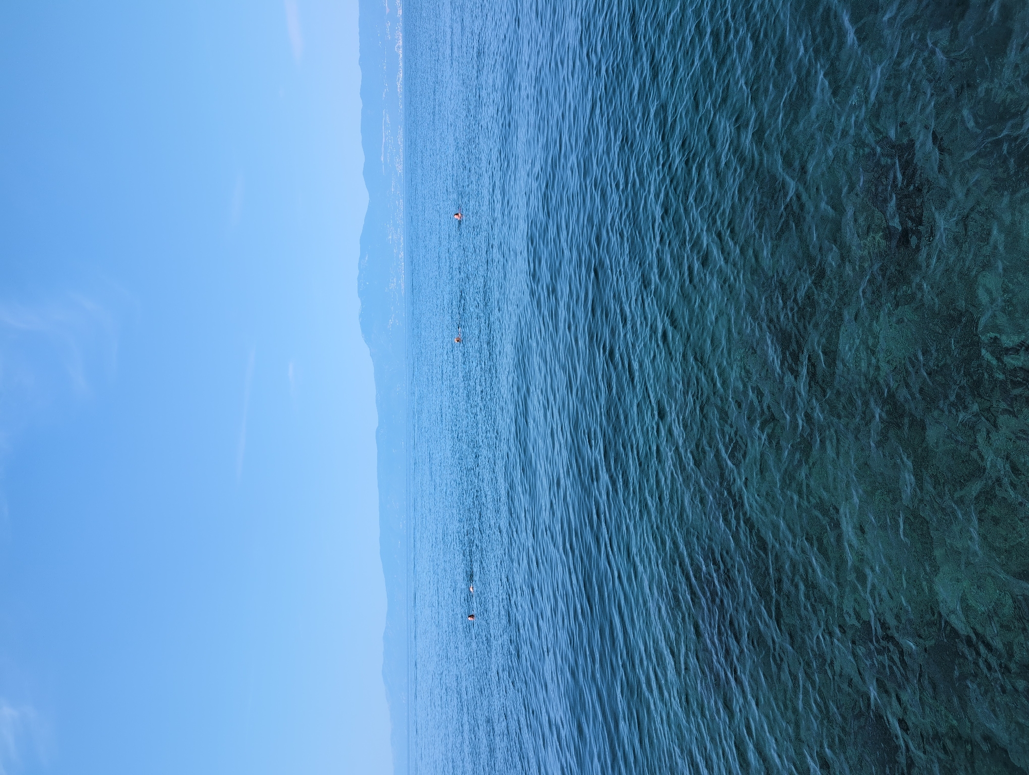 In Kostrena, Croatia, a tranquil seascape unfolds under a clear blue sky. The foreground is dominated by the shimmering turquoise water of the Adriatic Sea, its surface gently rippled, revealing the rocky seabed beneath. In the mid-distance, a few individuals are enjoying a swim, their heads and shoulders visible above the water. Further out, a hazy, mountainous coastline stretches across the horizon, punctuated by hints of urban settlements. The scene evokes a sense of peaceful recreation in a natural setting.
