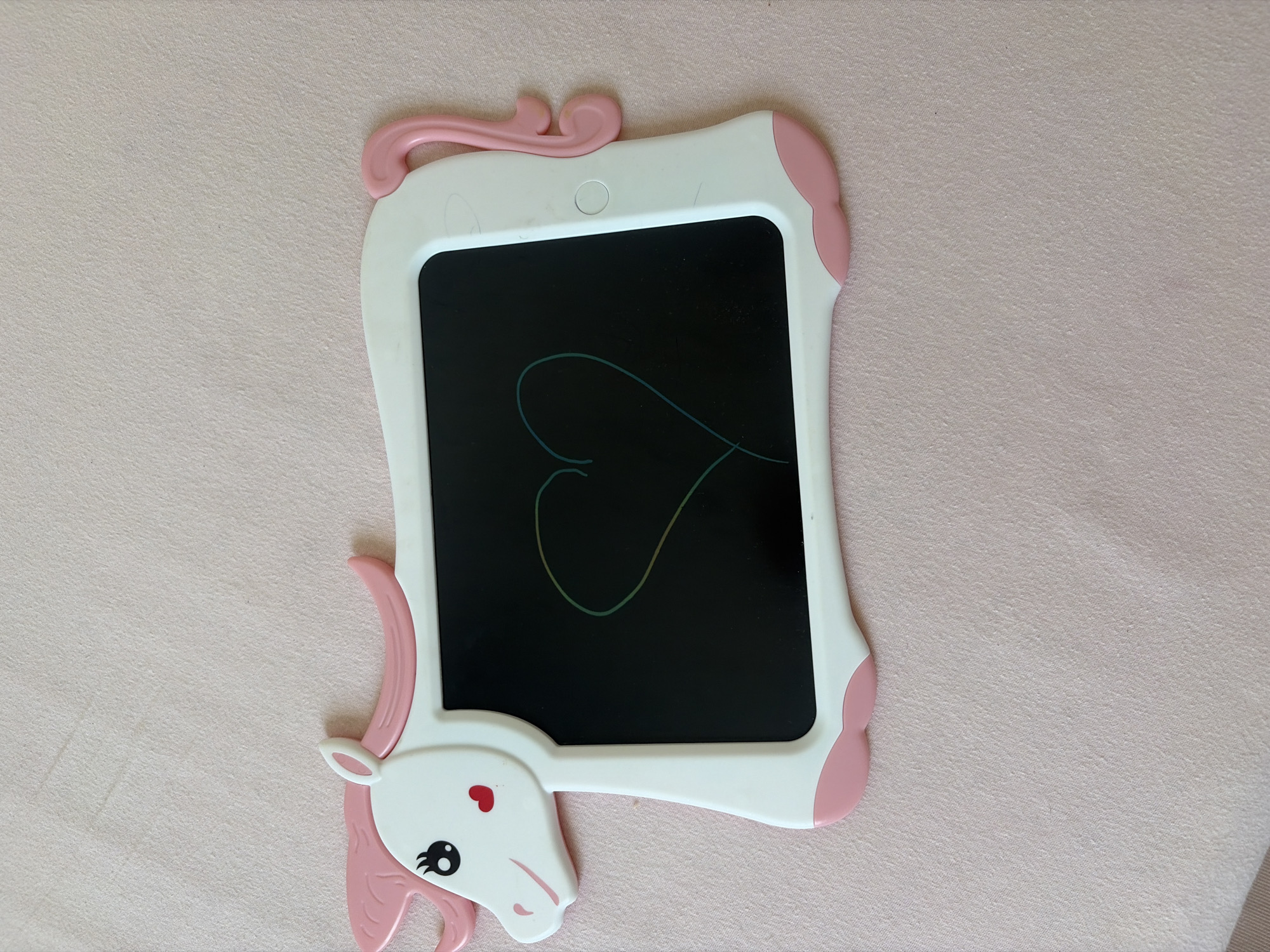 The image shows a pink and white unicorn-shaped digital drawing tablet resting on a pink, textured surface. The tablet has a black screen on which a faint, multi-colored heart shape has been drawn. The unicorn features include pink ears, mane, and tail, and a white head with black eyes and a small red heart on its cheek. There is no indication of a location in Duisburg, Germany, or any people present in the image.