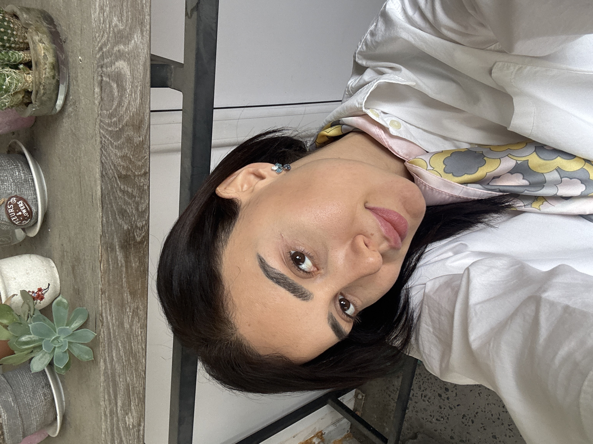 A woman with dark hair and brown eyes is facing the camera. She is wearing a white button-down shirt and a scarf with a pattern of yellow, gray, and pink. Her left ear is visible, adorned with a blue earring.
To the left of the woman, there is a wooden shelf with various plants in pots. There is a cactus, a succulent, and other greenery.
The background is a neutral wall with a glimpse of a metal structure, possibly a frame or scaffolding. The location of the image is not apparent from this image. It is not possible to determine whether this image was taken in Tbilisi, Georgia.