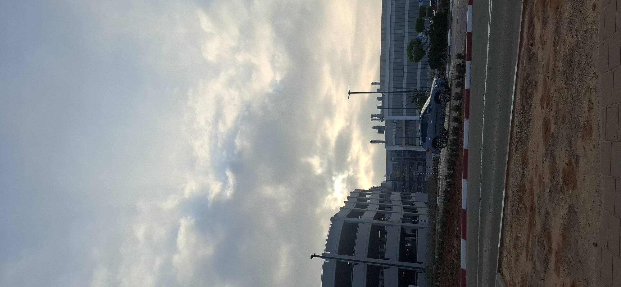 The image captures a street-level view in Kiryat Gat, Israel, during a cloudy day. In the foreground, a paved area with red and white borders separates the street from a strip of vegetation and then a road. A grey car is parked on the side of the road, facing right. Behind the car, a multi-story parking structure with grey concrete levels is visible on the left. Further in the background, a large industrial or commercial building complex with a glass facade and industrial chimneys is prominent. The sky is overcast with soft, diffused light filtering through the clouds, casting a gentle illumination on the scene. The overall impression is one of an industrial or business district with its characteristic structures and infrastructure.