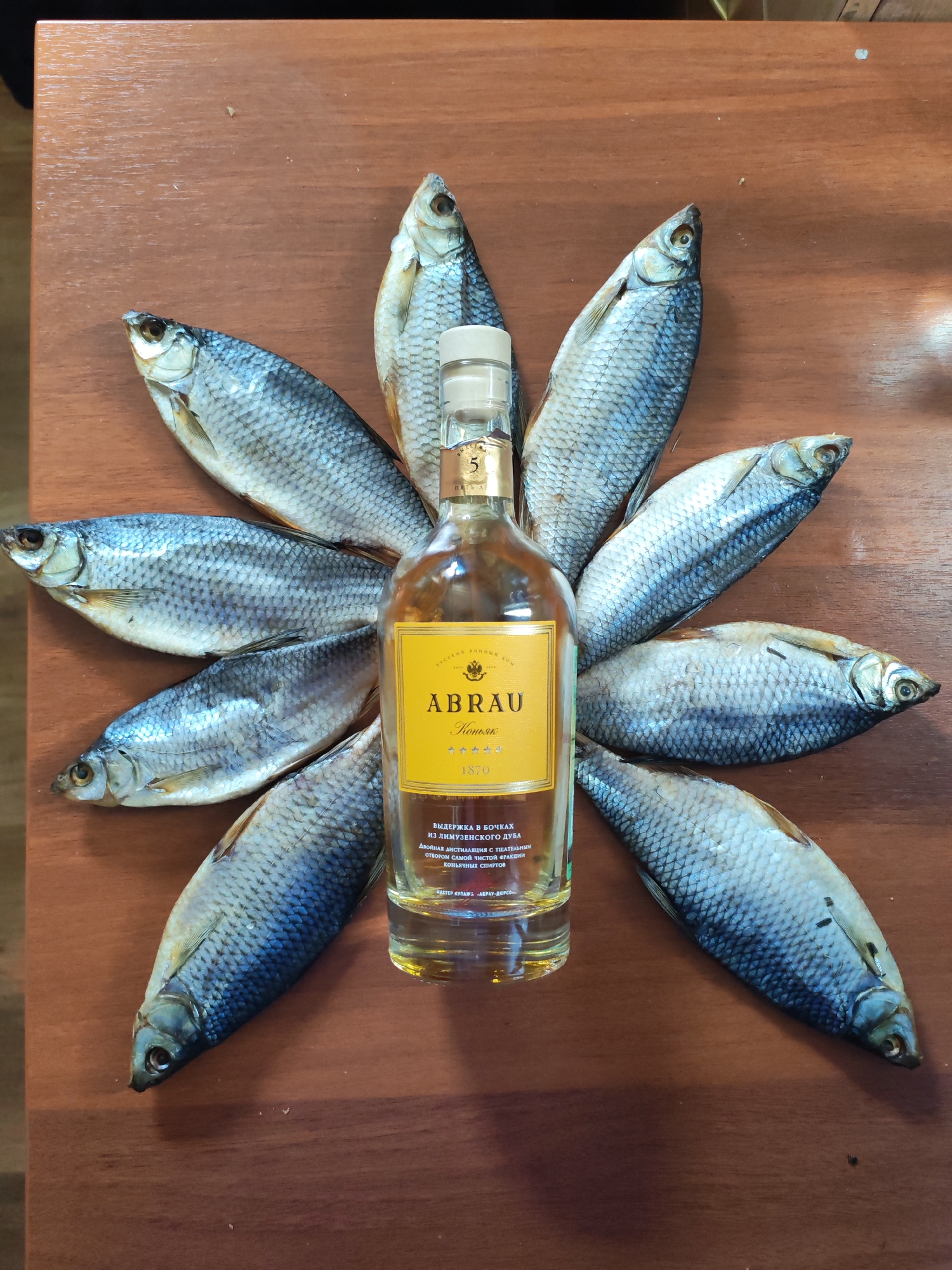 The image displays a still life arrangement on a polished, reddish-brown wooden table. At the center is a clear glass bottle of amber-colored brandy (cognac) with a yellow label. Surrounding the bottle, eight dried or cured fish are meticulously arranged in a radial, flower-like pattern, with their heads pointing outwards. The fish are uniformly sized, exhibiting visible scales and a silvery-grey hue.
The bottle's label prominently features the brand name "ABRAU" and the word "Коньяк" (Cognac) in Cyrillic. Below this, the year "1870" is inscribed. Further details on the label, translated from Russian, state "ВЫДЕРЖКА В БОЧКАХ ИЗ ЛИМУЗЕНСКОГО ДУБА" (Aged in Limousin oak barrels) and "ДВОЙНАЯ ДИСТИЛЛЯЦИЯ С ТЩАТЕЛЬНЫМ ОТБОРОМ САМОЙ ЧИСТОЙ ФРАКЦИИ КОНЬЯЧНЫХ СПИРТОВ" (Double distillation with careful selection of the purest fraction of cognac spirits). The bottle cap is white with the number "5" visible.
The scene captures a static display, with no people or active event taking place. The combination of brandy and dried fish suggests a traditional pairing, often enjoyed as a snack or accompaniment to alcoholic beverages in Russian culture, fitting the provided location context of Shchetinskoe, Russia.
