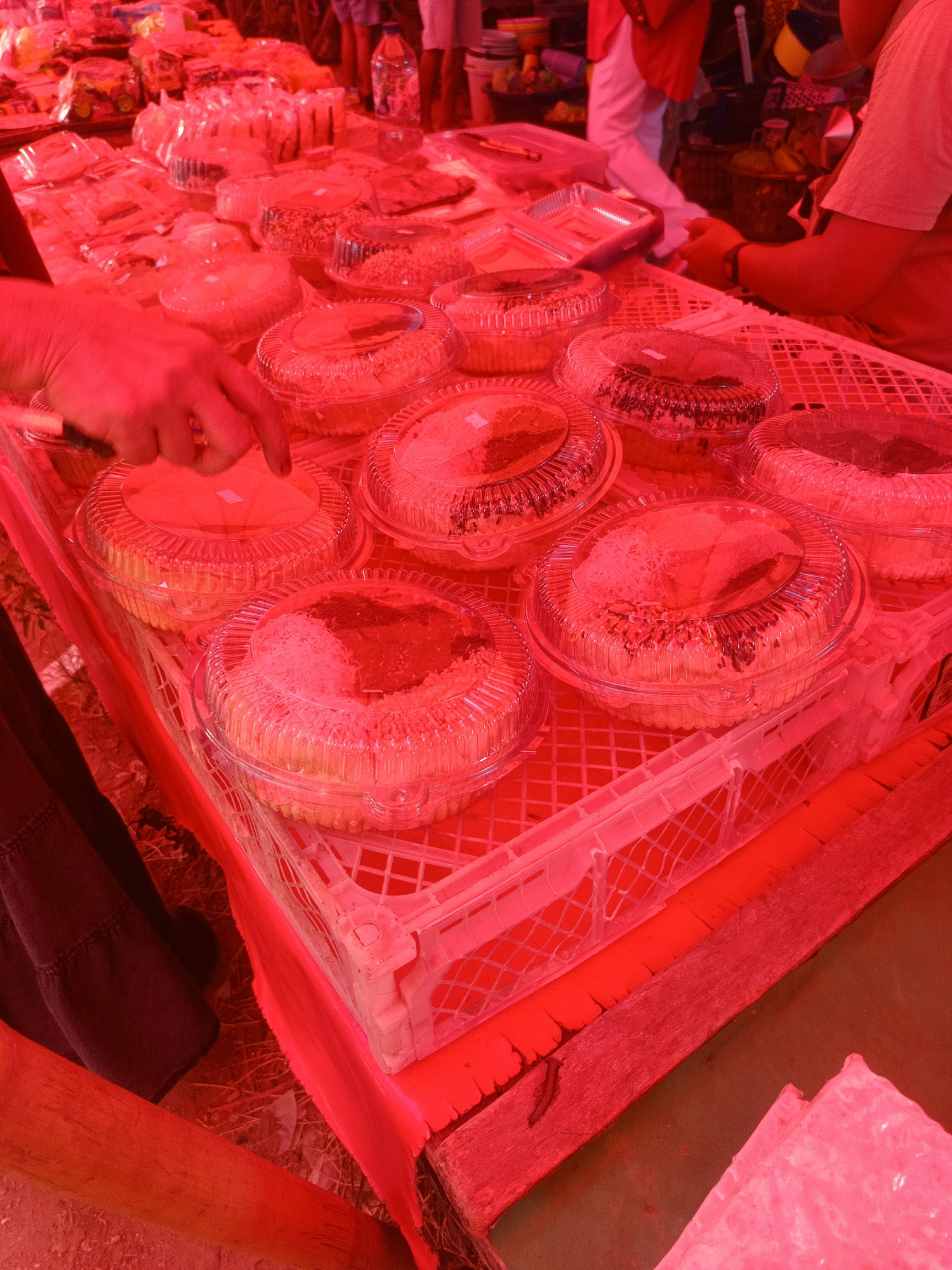 A low-angle, close-up shot captures a market stall in Gorontalo, Indonesia, dominated by a vibrant red hue due to intense lighting. The scene is filled with numerous transparent plastic containers, each holding baked goods, likely cakes or pastries. These containers are arranged on a white plastic crate, which sits on a wooden surface. A hand, illuminated in red, is visible reaching towards one of the containers, suggesting a transaction or selection. In the background, out of focus, are hints of people and other stalls, indicative of a bustling market environment. The overall impression is one of abundance and a lively commercial setting, albeit viewed through a strong red filter.