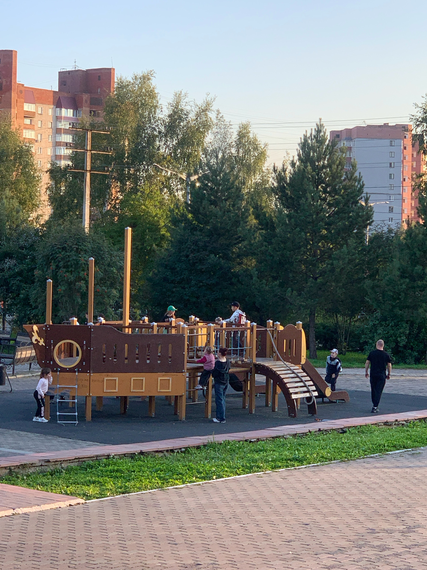 In Prokopyevsk, Russia, a playground featuring a pirate ship-themed climbing structure is the central focus. Several children are engaged in play on and around the structure, with some climbing, standing, and observing. A young girl in a pink vest is on the right side of the ship, while another girl in a white shirt and black leggings stands by a ladder on the left. A boy wearing a blue and grey jacket is near the slide, and another boy in a green hat is on the upper level of the ship. An adult male in a black t-shirt and pants walks away from the playground on the right side of the image. The playground is surrounded by trees and green grass, with apartment buildings visible in the background. The ground consists of a dark rubberized surface around the playground and brick paving in the foreground. The scene is illuminated by natural sunlight, suggesting it is daytime.