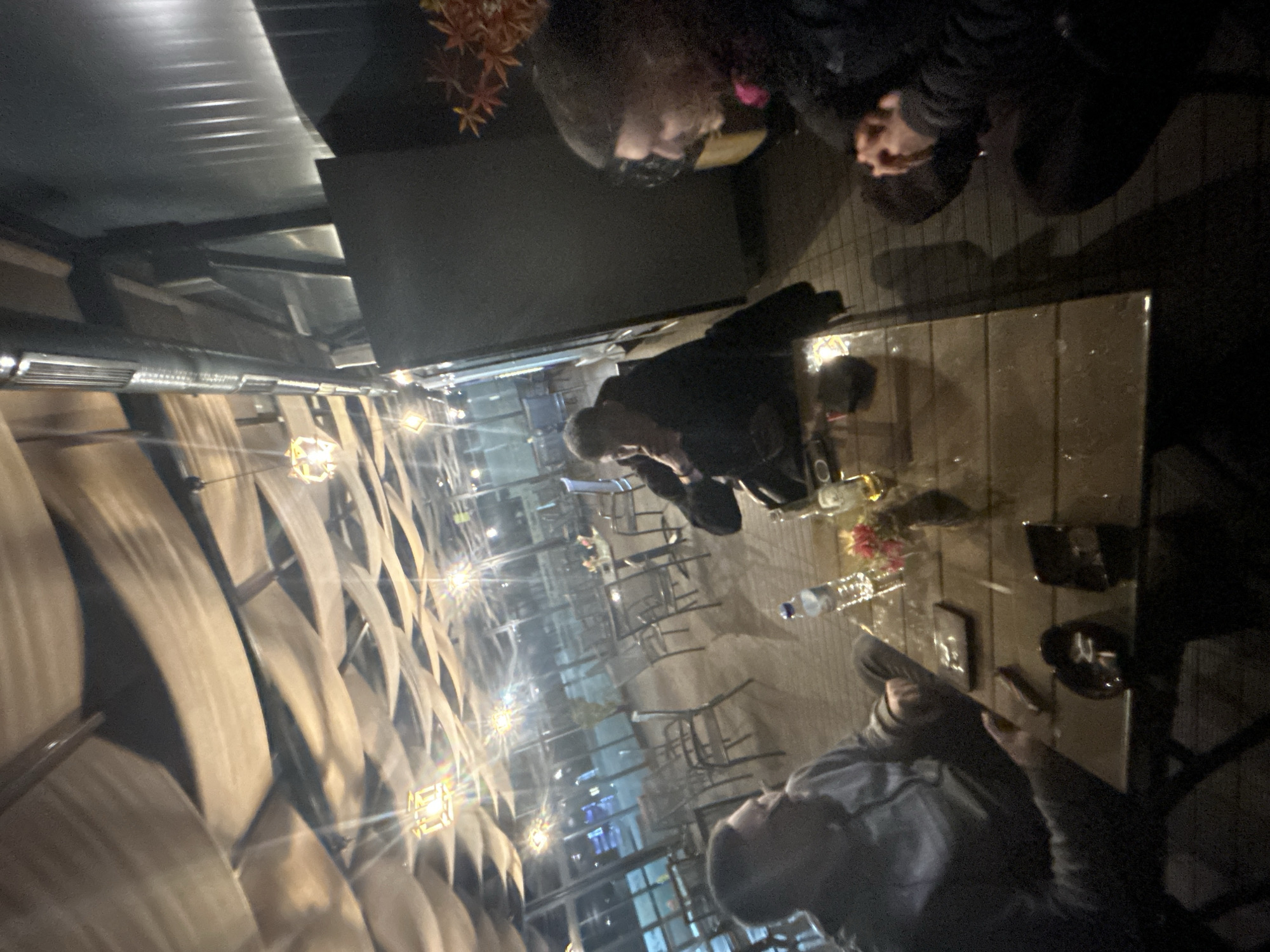 The image captures a scene within a covered outdoor seating area. Several people are seated at tables, engaged in conversation and presumably dining or socializing. The tables hold various objects, including bottles, glassware, and what appear to be decorative floral arrangements. The ceiling of the seating area has a textured, woven design illuminated by multiple light fixtures. Reflections in the glass surfaces of the tables indicate the presence of windows and other light sources. There is no other visual information to suggest location.
