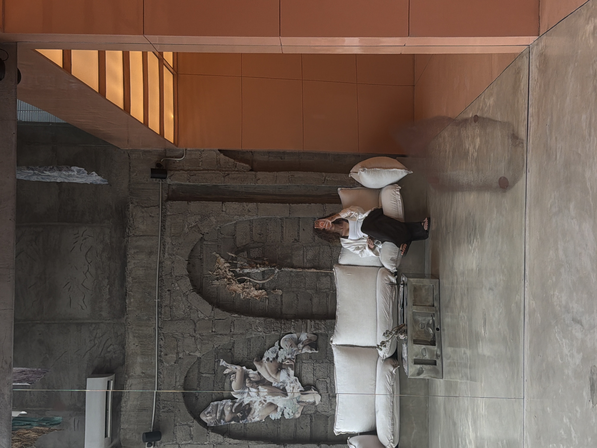 The image captures a modern, minimalist interior space with a rustic industrial aesthetic, likely located in North Kuta, Indonesia. A large, off-white sectional sofa dominates the foreground, positioned against a textured grey stone wall. The wall features two arched recesses, one containing a collage-like artwork depicting figures and the other displaying a dry, branch-like plant. A sleek, metallic coffee table sits in front of the sofa, adorned with a small decorative branch.
A woman with curly brown hair is seated on the right side of the sofa, holding a smartphone to take a selfie. She is wearing a white tank top and dark trousers, with her legs crossed. Beside her is a water bottle.
The flooring is polished concrete, reflecting the overhead lighting and the surroundings. To the right of the stone wall, a section of the space is illuminated by a warm, orange-toned light emanating from a ceiling structure composed of horizontal panels. This area is clad in smooth, matte orange panels.
A visible reflection on the polished concrete floor shows the blurred outline of what appears to be a person and furniture, suggesting the presence of a reflective surface, likely glass, in front of the scene. An air conditioning unit is mounted on the grey wall above the sofa. The overall impression is one of a chic, contemporary lounge or café, possibly a popular spot in North Kuta for relaxation and social media engagement.