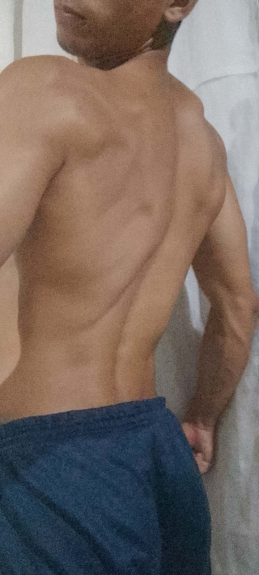 A man stands with his back facing the camera, showcasing defined back muscles and arm muscles. He is wearing blue shorts. The background appears to be a plain wall, possibly white or light-colored. The image quality is slightly grainy. The location in Juan Griego, Venezuela is not discernible from the image.