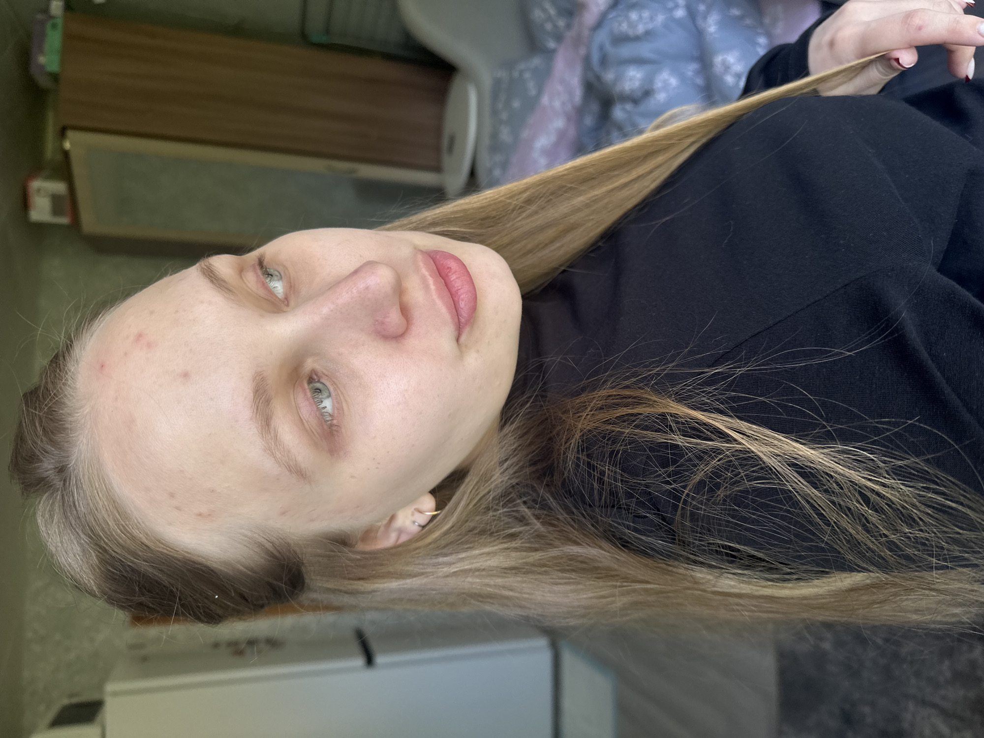 A young, fair-skinned person with light blonde hair and green eyes is facing the camera. They appear to be wearing a black turtleneck sweater. The person's skin exhibits some minor blemishes. The background is blurry and indistinct, containing elements of what could be furniture. It is impossible to determine the specific location in Chaikovsky, Russia, or confirm it is even in Chaikovsky.