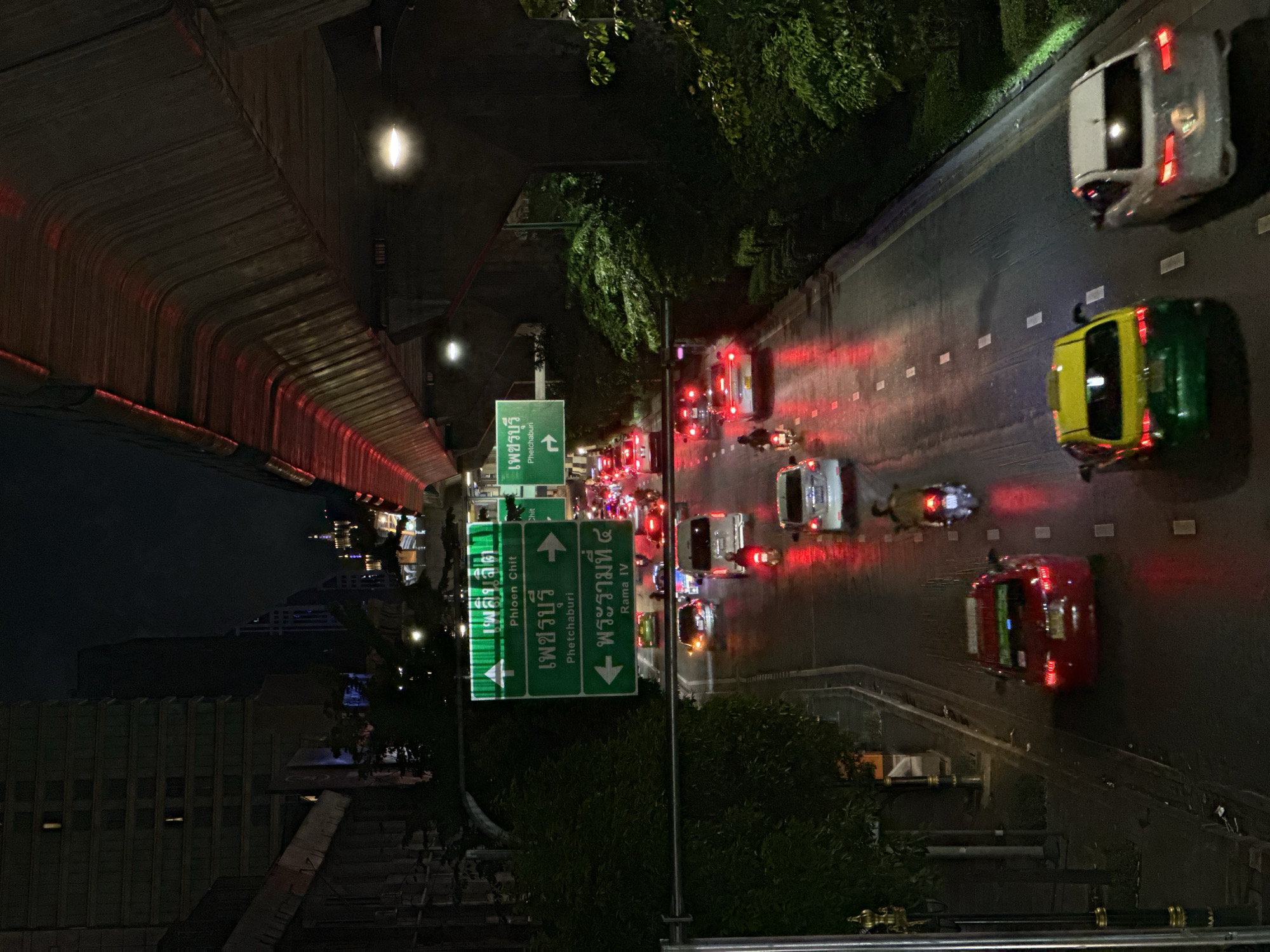 The image depicts an aerial view of a street in Bangkok, Thailand at night. A road sign with directions to "Petchaburi" and "Rama IV" indicates the location. A bridge or elevated structure runs along the left side of the image, casting a shadow over a building. Traffic is visible on the road, including cars, taxis, and motorcycles. The taillights of the vehicles create streaks of red light. Trees are present along the side of the road, partially obscuring the view.