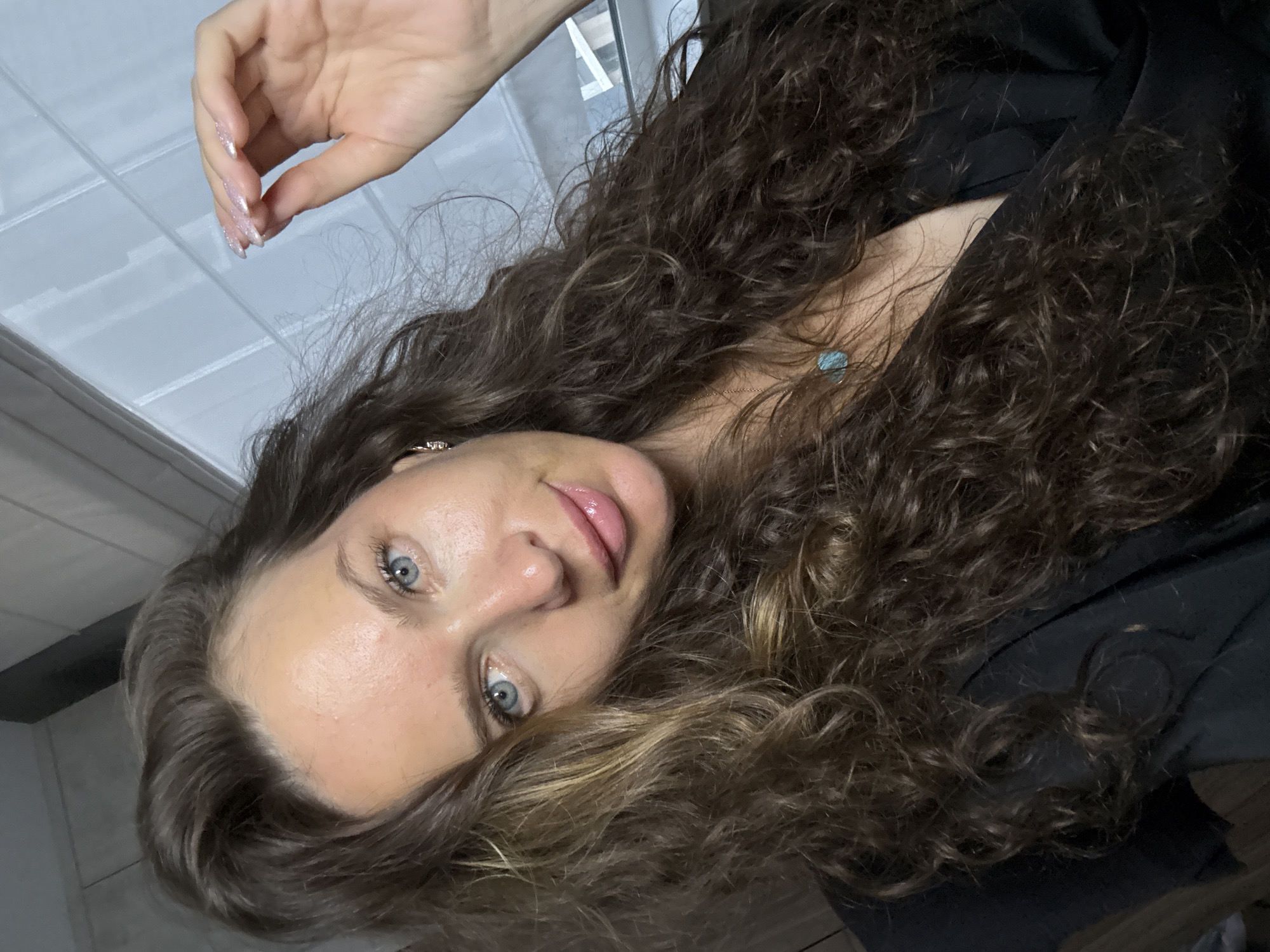 A woman with long, wavy brown hair is shown in a close-up, selfie-style portrait. She has blue eyes and is wearing a black top and a necklace with a turquoise pendant. Her right hand is raised towards her hair, and her nails appear to be painted with a glittery polish. The background is out of focus, but suggests an indoor setting with natural light coming from a window with blinds. There is no information provided to determine her location in Moscow, Russia.