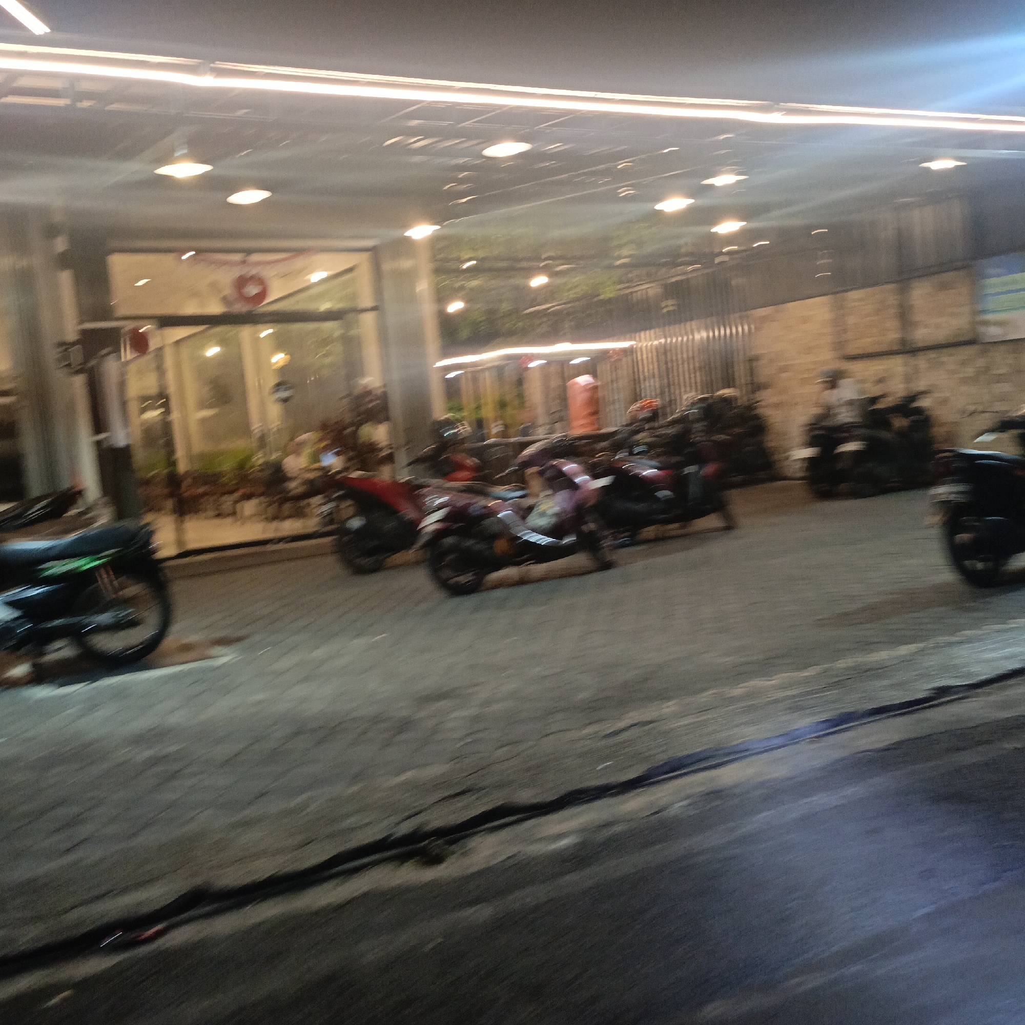 The image depicts a nighttime scene outside a building with large windows and several parked motorcycles. The building appears to be a commercial establishment, possibly a cafe or restaurant, indicated by the interior lighting and seating visible through the windows. The exterior is lit by overhead lights, including some strip lighting along the ceiling and individual pendant lights. The ground is paved with interlocking bricks, common in urban outdoor spaces in Indonesia. Several motorcycles are parked in a row, some with riders, suggesting it is a popular gathering spot. One motorcycle in the foreground, on the left, is black with green accents. Other motorcycles are red and dark colored. The overall impression is one of a bustling urban environment at night. The specific location in Salatiga, Indonesia, is not discernible from the image alone, but the scene is consistent with many commercial areas in Indonesian cities.