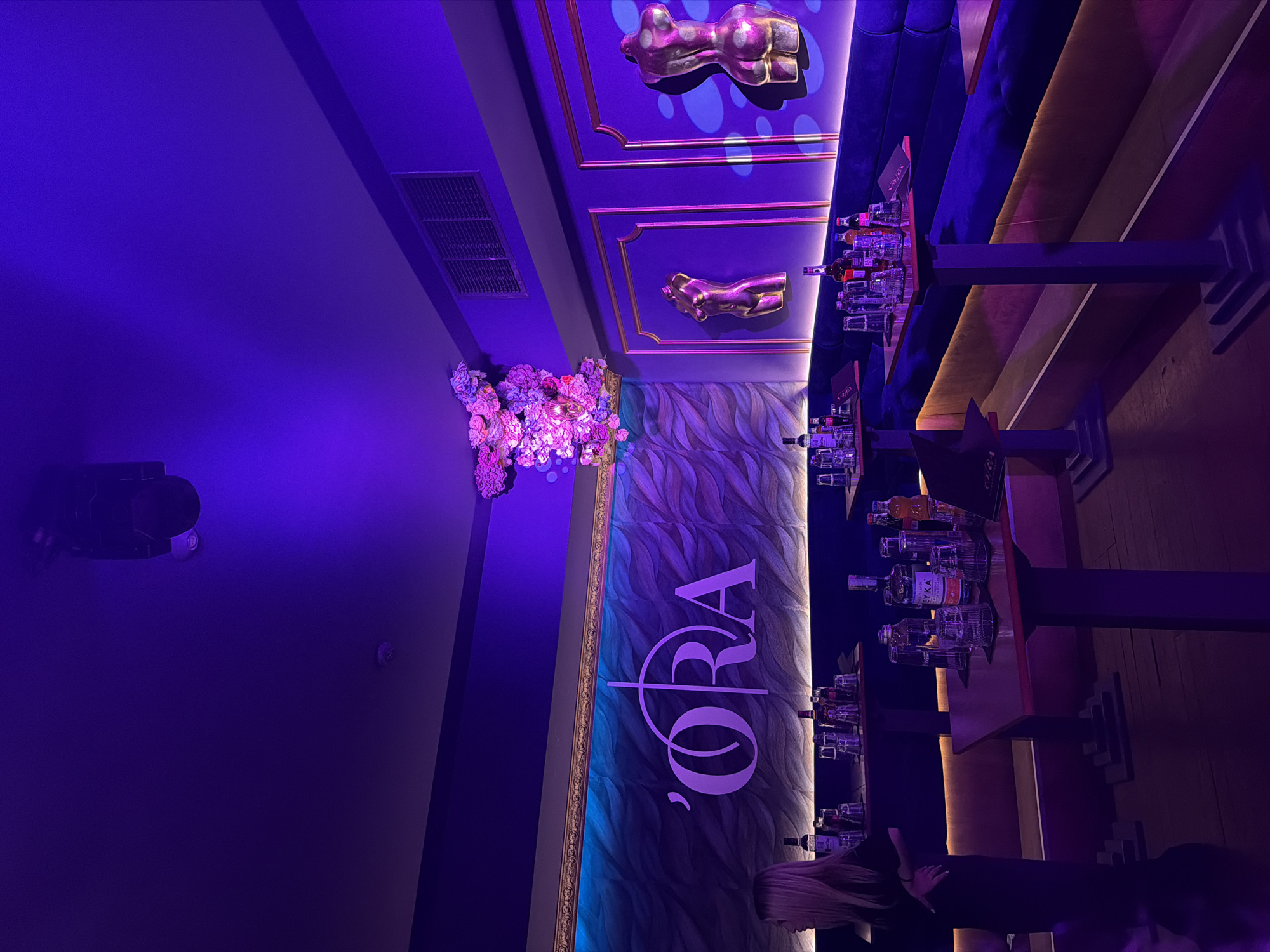 The image shows the interior of a bar or lounge, possibly in Blagoevgrad, Bulgaria, featuring a dark, purple-lit environment. Prominently displayed is the bar's name, "Ora," in large white lettering against a textured wall with wave-like patterns. The bar counter is stocked with bottles and glasses, arranged on shelves. The ceiling has decorative elements, including gold-colored body sculptures and floral arrangements. The lighting is primarily purple, creating a dramatic atmosphere. A person with blonde hair is partially visible near the bar.