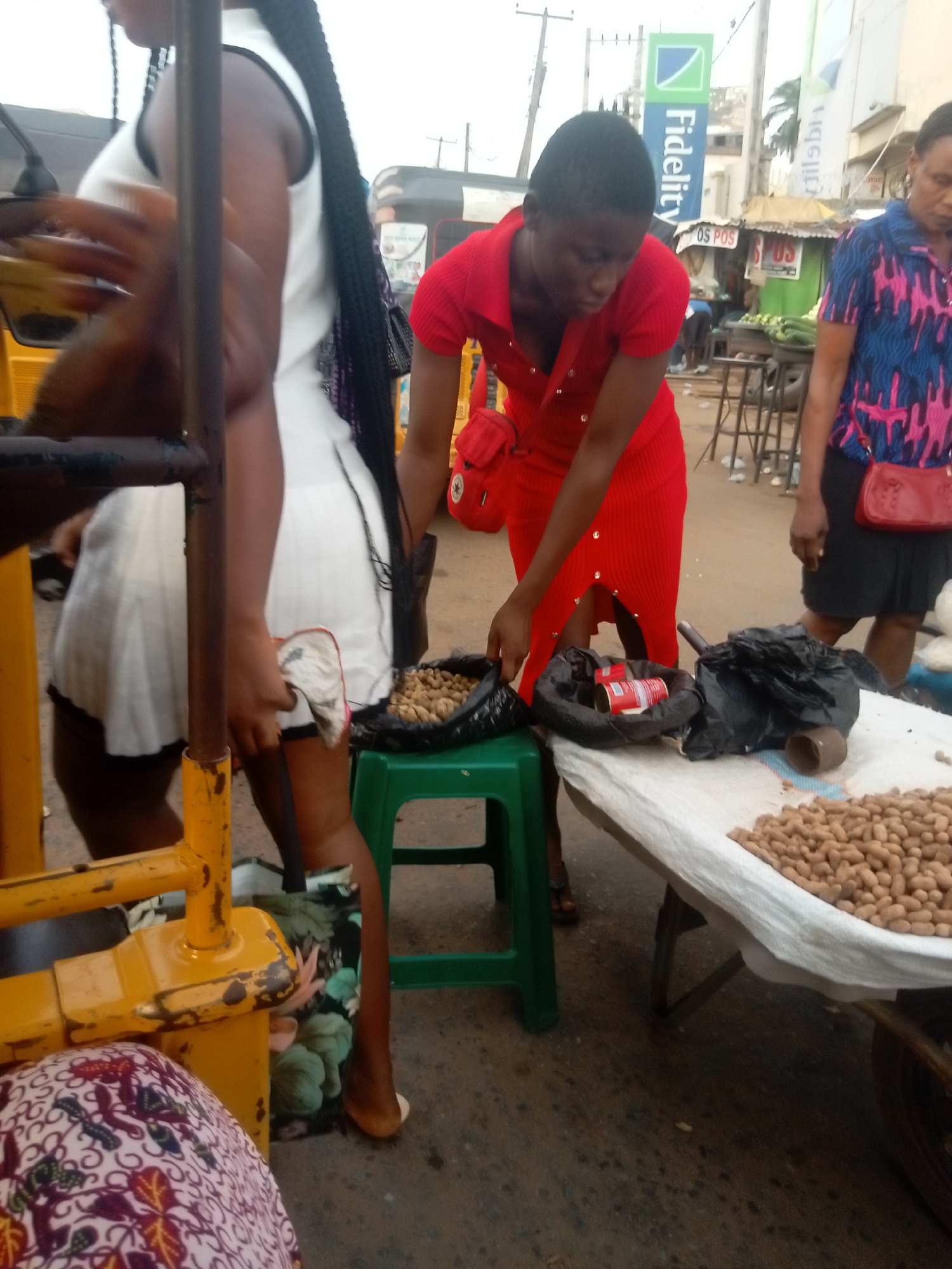 The image depicts a street scene in Nkpor, Nigeria. A woman in a red dress is bending over a table covered with a white cloth, handling peanuts. Some peanuts are spread on the table, while others are contained in a black bag on a green plastic stool. Another woman in a blue patterned shirt and black skirt stands beside the peanut table. Fidelity Bank signage is visible in the background. A woman wearing a white dress and black braids is standing to the left of the woman in red dress, standing at the yellow auto rickshaw parked on the street. The general environment appears to be a busy market area.