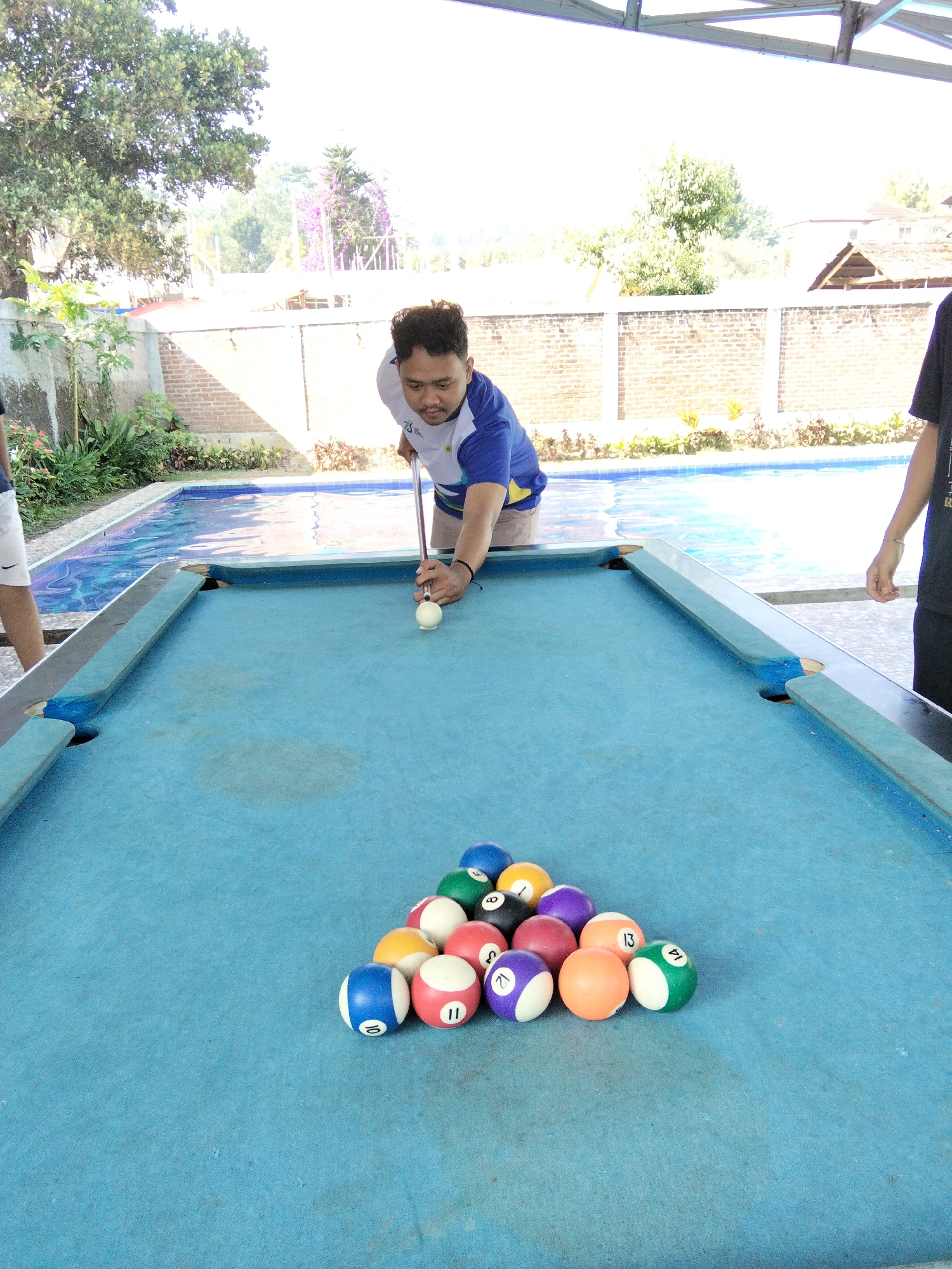 The image shows a man playing pool next to a swimming pool. The man is wearing a blue and white shirt and is holding a pool cue. He is about to strike a white ball on the pool table. Several numbered billiard balls are racked in a triangle on the table. The scene is outdoors, with a swimming pool, brick wall, and trees visible in the background. The architecture and vegetation suggest a location in Cisarua, Indonesia, likely a resort or private residence.