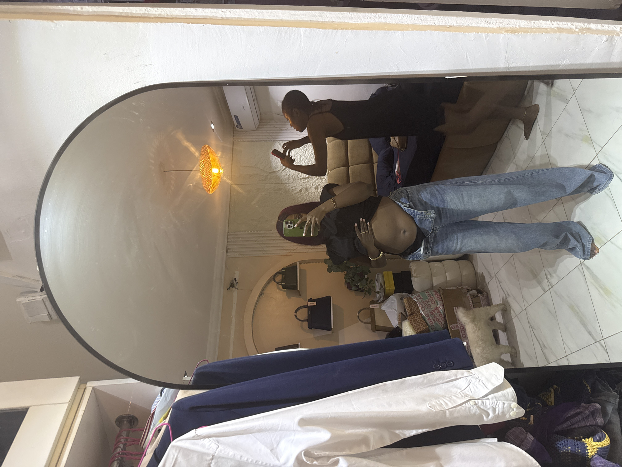 A young woman with dark hair, wearing a black crop top and blue jeans, stands in a bedroom and takes a mirror selfie. She is in the process of adjusting her jeans, which are unbuttoned at the top, exposing her midriff. Her phone, held in her right hand, is angled to capture her reflection. In the mirror's reflection, another young woman in a black slip dress is visible in the background, holding a red phone to her ear as if on a call. The bedroom features a large, tufted headboard, a bedside table with a bag, and a woven lampshade hanging from the ceiling. A small, light-colored dog is walking across the tiled floor in the foreground, near a closet filled with clothes. The room appears to be located in a residential dwelling in Abuja, Nigeria.