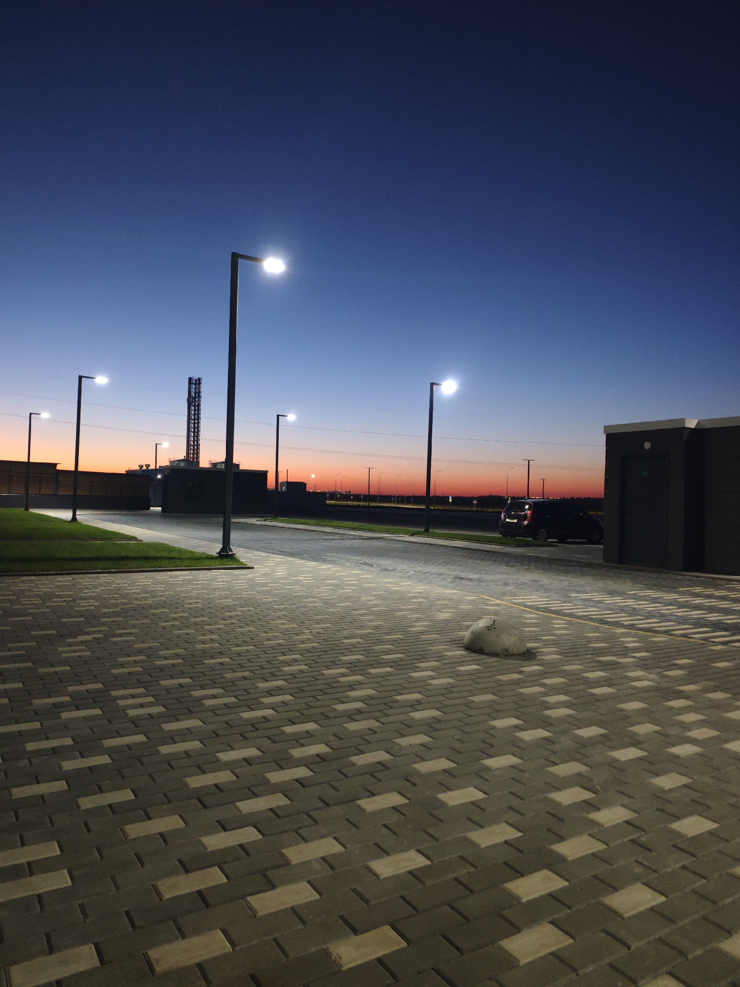 The image depicts an outdoor scene at dusk, likely in an industrial or commercial area in Krasnodar, Russia. The sky transitions from a deep blue at the top to a vibrant orange and red gradient near the horizon, indicating the setting sun. Several modern streetlights with illuminated bulbs line a paved pathway, casting a glow on the interlocking gray and tan brickwork.
To the left, a well-maintained lawn borders the paved area, with a short fence or wall behind it. Further back, a dark building with a tall industrial structure, possibly a chimney or tower, rises against the colorful sky. A dark-colored SUV is parked on the road or adjacent area to the right of the frame.
In the foreground, a small, light-colored object, appearing to be a dog or a large stuffed animal, rests on the brick pavement. To the far right, a modern, dark-colored structure with a door is visible, possibly a utility building or storage unit. The overall scene is quiet and still, with the artificial lights of the streetlamps beginning to dominate as daylight fades.