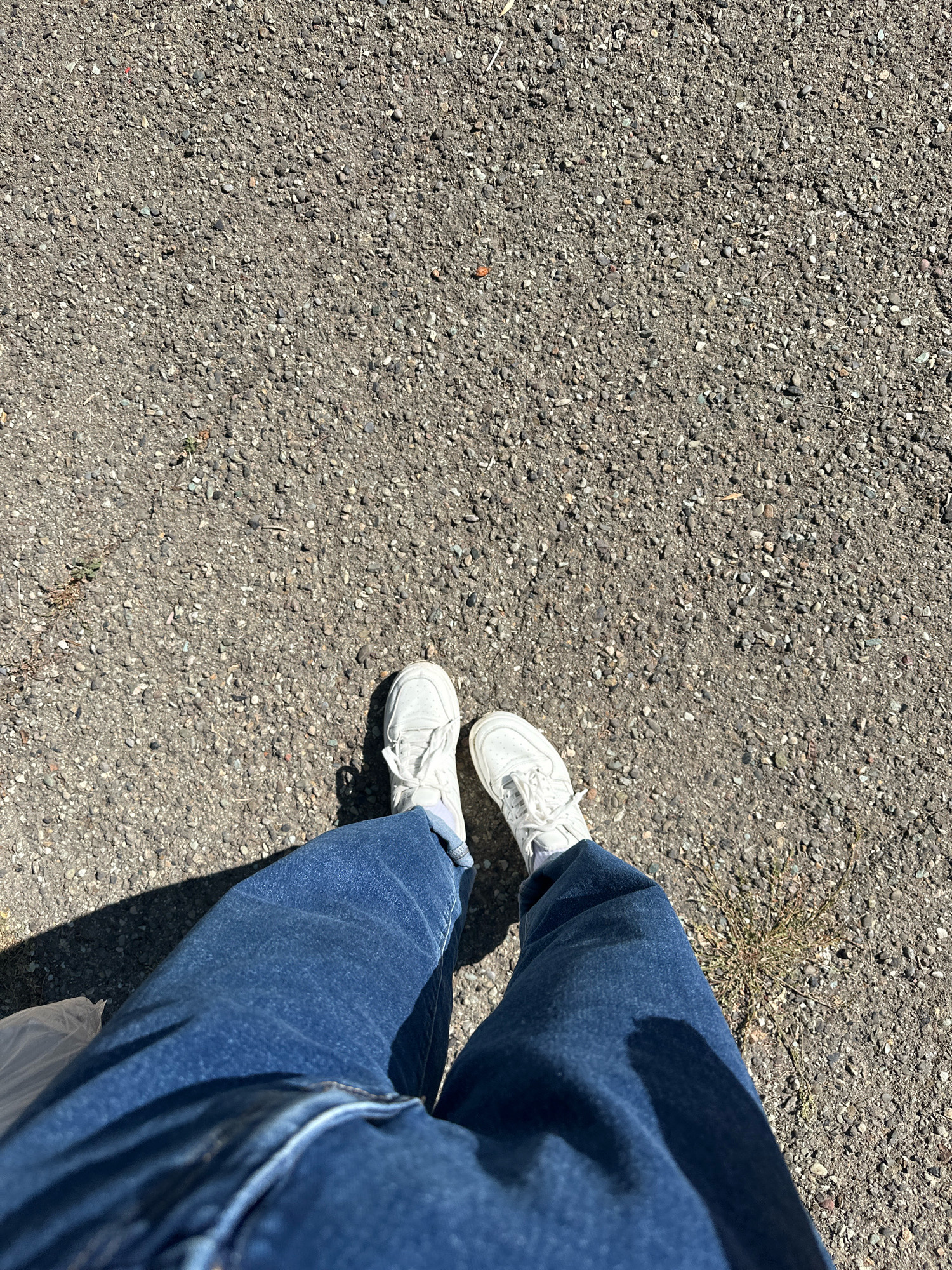 This image depicts a top-down view of a person's legs and feet standing on an asphalt surface. The asphalt is dark gray and composed of small pebbles. The person is wearing blue denim jeans and white athletic shoes with white socks. The lighting suggests it is daytime, with a shadow cast from the person to their left. The environment appears to be an outdoor area, possibly a street or walkway in Gyumri, Armenia, given the context. There are no other people or significant objects in the frame besides the person's attire and the ground.