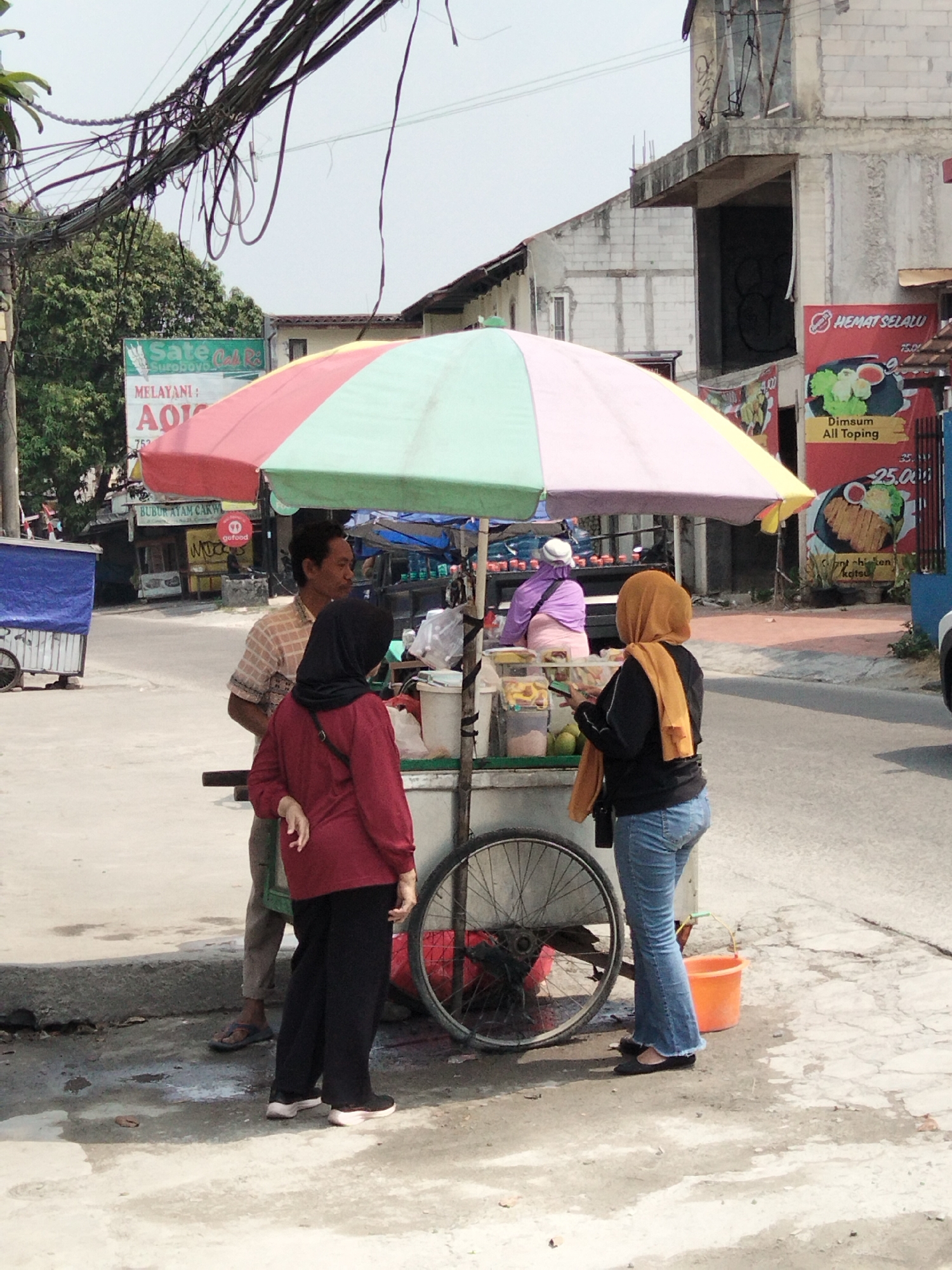 This is a street scene photograph taken in Depok, Indonesia, depicting a food cart on a paved sidewalk. A colorful umbrella, segmented in red, yellow, green, and pink, shades the cart and three individuals.
In the foreground, a person wearing a black hijab, orange scarf, black jacket, and blue flared jeans stands facing away from the camera, holding a phone. To their left, a person in a dark red top and black pants, also wearing a black hijab, stands with their back partially to the camera. To the left of these two, a man in a checkered shirt and dark pants stands facing the cart. The food cart itself has a wheel visible on the right side, and displays various food items and containers.
In the background, a building with exposed brickwork and unfinished construction is visible. A red banner with text and images of food, possibly advertising "Dimsum All Toping," is attached to the building. Other street vendors and structures are present in the distance. Overhead, a dense network of electrical wires crisscrosses the sky. The ground is a mix of concrete and dirt, with some patches of water. The overall atmosphere suggests a bustling urban environment.