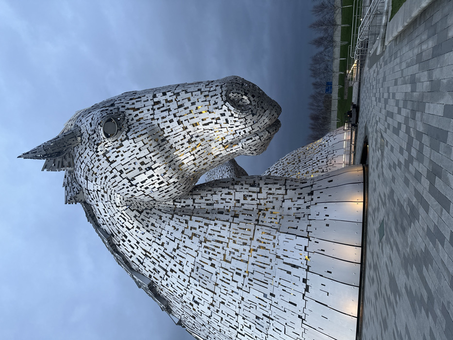 The image displays one of 'The Kelpies' monumental horse head sculptures, located in Helix Park, Falkirk, United Kingdom, under an overcast sky. The sculpture is constructed from numerous interlocking rectangular and irregular brushed metal panels, presenting a silver-grey appearance. Warm yellow light emanates from internal sources through gaps in the plating and from ground-level spotlights illuminating its base. The horse's head is angled downwards and to the right, featuring a distinct circular eye detail. The foreground consists of a grey paved pathway composed of rectangular pavers. In the midground, a grassy bank rises, populated by a line of leafless deciduous trees and bordered by a metal railing. No individuals are visible, and no discernible actions or interactions are occurring within the scene.
