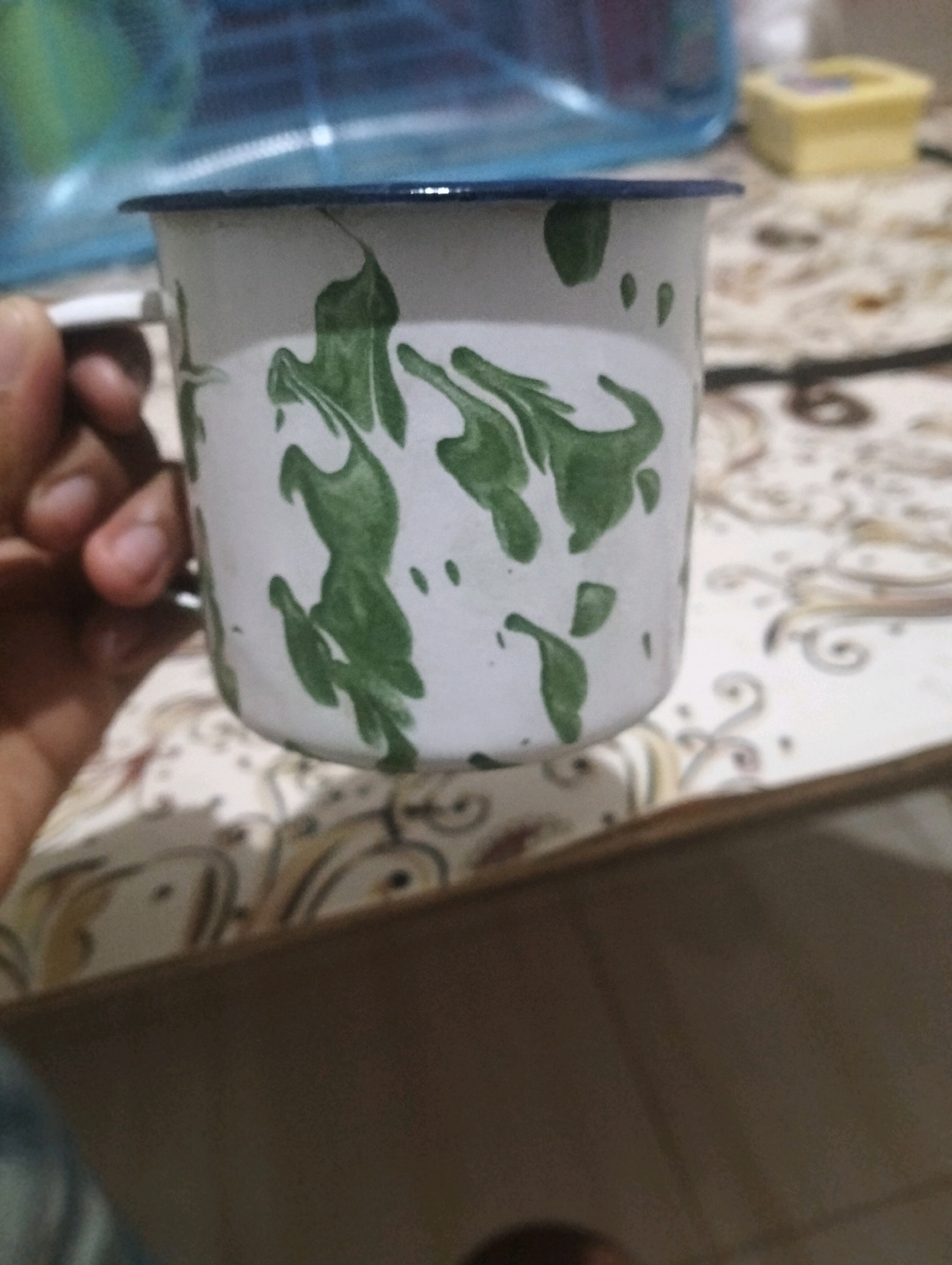 A close-up, eye-level, static shot captures a hand holding a white enamel mug with a blue rim and a green abstract pattern. The mug is positioned in the foreground. The background is blurred, showing a patterned tablecloth, a yellow object, and a blue translucent container. The scene appears to be indoors, possibly in a home in Klecorejo, Indonesia, based on the provided location information.