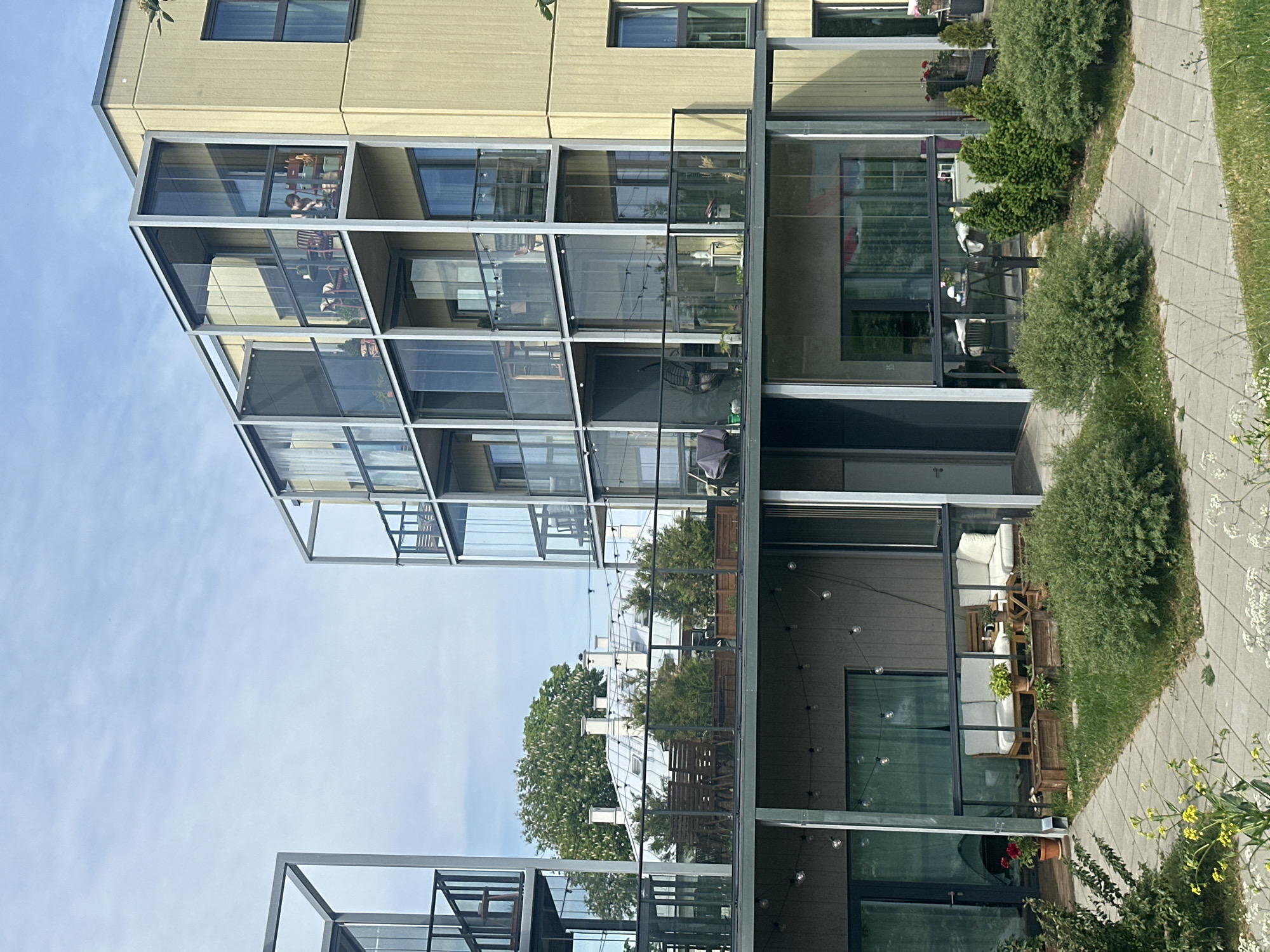 The image captures the exterior of modern apartment buildings in Tallinn, Estonia, featuring balconies with glass enclosures. In the foreground, a paved walkway runs through a grassy area with various green bushes and some yellow wildflowers. The buildings themselves are characterized by a pale yellow facade and metal framing for the balconies. On the ground level, a patio area is visible with outdoor furniture, including a white sofa set and a wooden coffee table, along with potted plants and string lights. Higher levels show more balconies, some furnished with chairs and tables, and one with a person visible through the glass. In the background, a building with a distinctive mansard roof and multiple chimneys can be seen, suggesting an older architectural style contrasting with the modern apartments. The sky is partly cloudy.