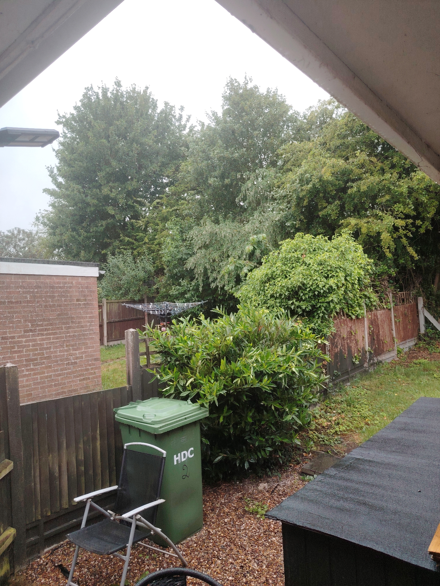 The image displays an outdoor residential scene, likely a garden or backyard, observed from a slightly elevated vantage point within Huntingdonshire, United Kingdom. The sky is heavily overcast and bright white, suggesting a dull or rainy day, with visible water droplets on some surfaces.
In the upper left and right, segments of a light-colored, angled architectural structure, possibly a roof overhang or beam, partially frame the view. A black, modern-style street light fixture is visible on the far left, partially obscured by this structure.
The background is dominated by a dense canopy of various deciduous trees with dark green foliage. Below these trees, a weathered wooden fence line is visible, with some sections appearing discolored and possibly covered in rust or overgrown vegetation. A detached, single-story brick building with a flat roof is situated on the left. The bricks are reddish-brown, and the flat roof has a light-colored fascia. To the right of the brick building, a folded or collapsed rotary clothes airer is visible beyond the fence line.
In the lower left and center, a green wheeled waste bin with a closed lid is positioned on a gravel surface. The letters "HDC" and the number "2" are printed in white on its side, identifying it as belonging to the Huntingdonshire District Council waste collection system. Adjacent to the bin, a black fabric and silver-framed folding garden chair is unfolded and resting on the gravel.
The immediate foreground on the right features the dark, flat roof of a shed or similar outbuilding, covered in a dark, textured roofing material, likely felt. The ground in the foreground consists of light brown and grey gravel. Various green bushes and shrubs with dense foliage are scattered throughout the midground and foreground, bordering the fences and gravel areas. No individuals are present in the scene.