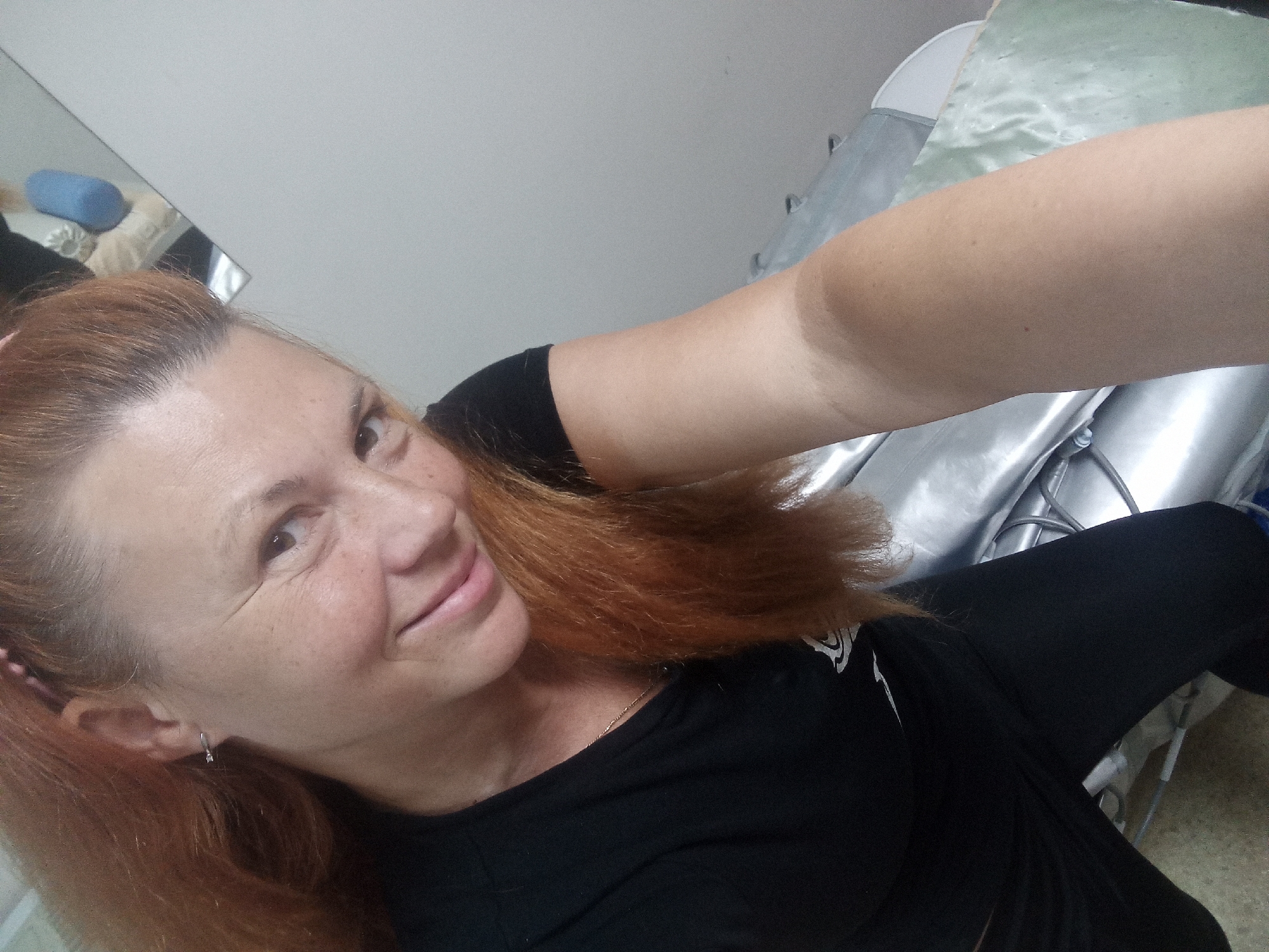 A woman with auburn hair is taking a selfie in what appears to be an indoor setting. She is wearing a black t-shirt and black leggings. Her hair is down and appears to be somewhat wind-swept. She has freckles on her face and is smiling. In the background, there is a mirror reflecting some objects, including a blue cylindrical object and a tan cloth item. To the right of the woman, there is a shiny, silver surface, possibly a piece of furniture or equipment, with some metal supports. The lighting suggests it might be daytime, but there are no direct indications of outdoor weather. The overall impression is a casual, personal moment captured by the subject.
