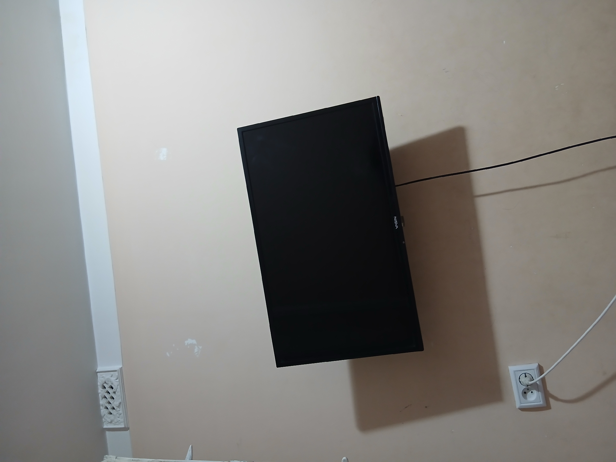 A wall-mounted black television screen with the brand name "VASIN" visible on its bottom edge is angled on a cream-colored wall in Margilan City, Uzbekistan. To the left of the television, near the top of the wall, is a white air vent with a geometric pattern. Below the television and to the left, a white electrical outlet with two circular sockets is visible, with a white power cord plugged into one of them and extending downwards and to the right. The wall has some minor imperfections and scuff marks. The lighting suggests an indoor setting.