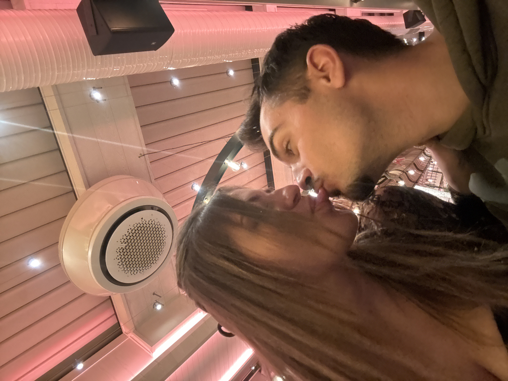 The image captures two individuals, a female and a male, engaged in an intimate interaction within an indoor commercial establishment in the city of Sofia, Bulgaria. Both subjects are positioned in profile, facing each other from the chest up, with puckered lips and extended tongues, their faces in close proximity. The female, on the left, has long, light brown hair that partially obscures her face and appears to be wearing a dark, possibly patterned, top. The male, on the right, has short dark hair, light facial stubble, and is wearing a dark green hooded sweatshirt.
The background features a modern ceiling composed of light-colored panels. The environment is illuminated by warm, reddish-pink ambient light emanating from concealed strip lighting, supplemented by several circular spotlights. Overhead, a prominent white, circular ceiling-mounted air conditioning unit with a black rim and patterned grille is visible. To the right, a corrugated white ventilation duct and a dark, rectangular speaker unit are mounted near the ceiling. The overall setting is consistent with a modern bar, club, or restaurant.