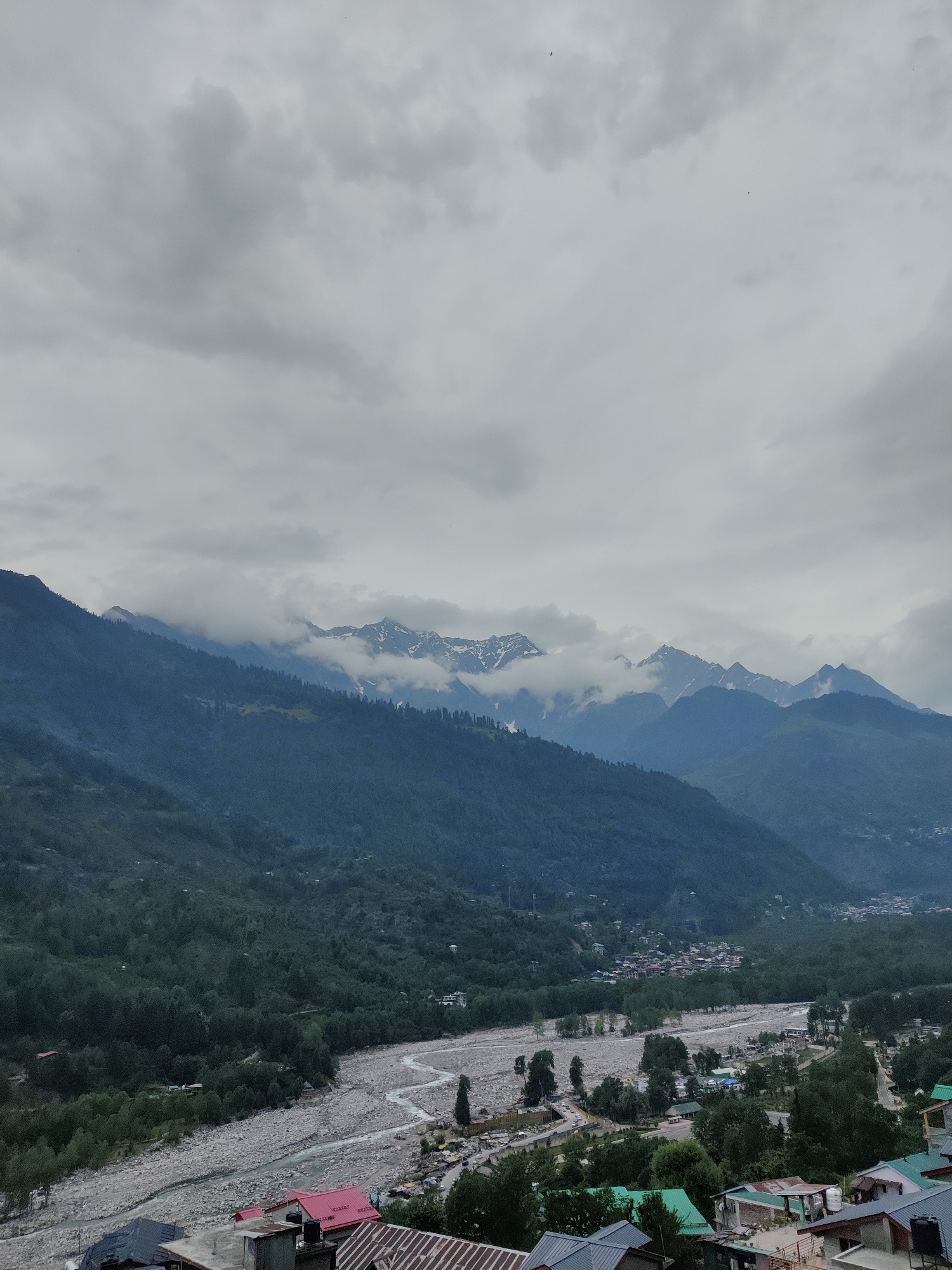 The image depicts a panoramic view of a valley in Vashisht, India, under a cloudy sky. The foreground features rooftops of buildings with red, green, and grey shingles, suggesting a residential or tourist area. A wide, stony riverbed cuts through the middle ground, with a winding river flowing through it. Lush green trees and dense vegetation cover the slopes of the mountains that dominate the background. The mountains are layered, with the closest ones appearing dark green and densely forested, while the further ones are capped with snow and shrouded in clouds, creating a dramatic and majestic landscape. A small town or village is nestled in the valley, visible between the river and the forested slopes. The overall impression is one of natural beauty and a remote, mountainous setting.