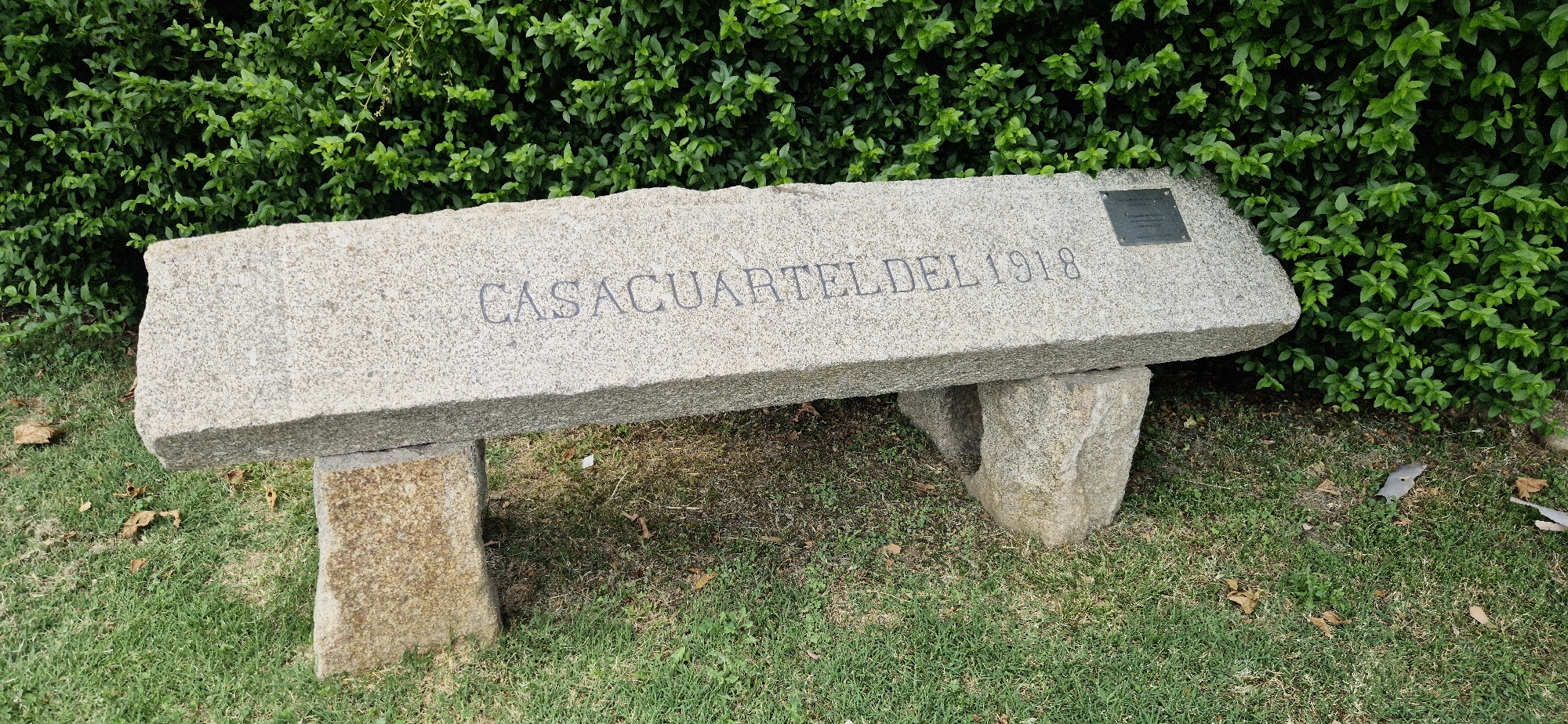 A stone bench with the inscription "CASACUARTEL DEL 1918" is situated in front of a dense green hedge. The bench is supported by two rough-hewn stone blocks. A small plaque is affixed to the right side of the bench. The ground beneath and around the bench is covered in grass and fallen leaves.