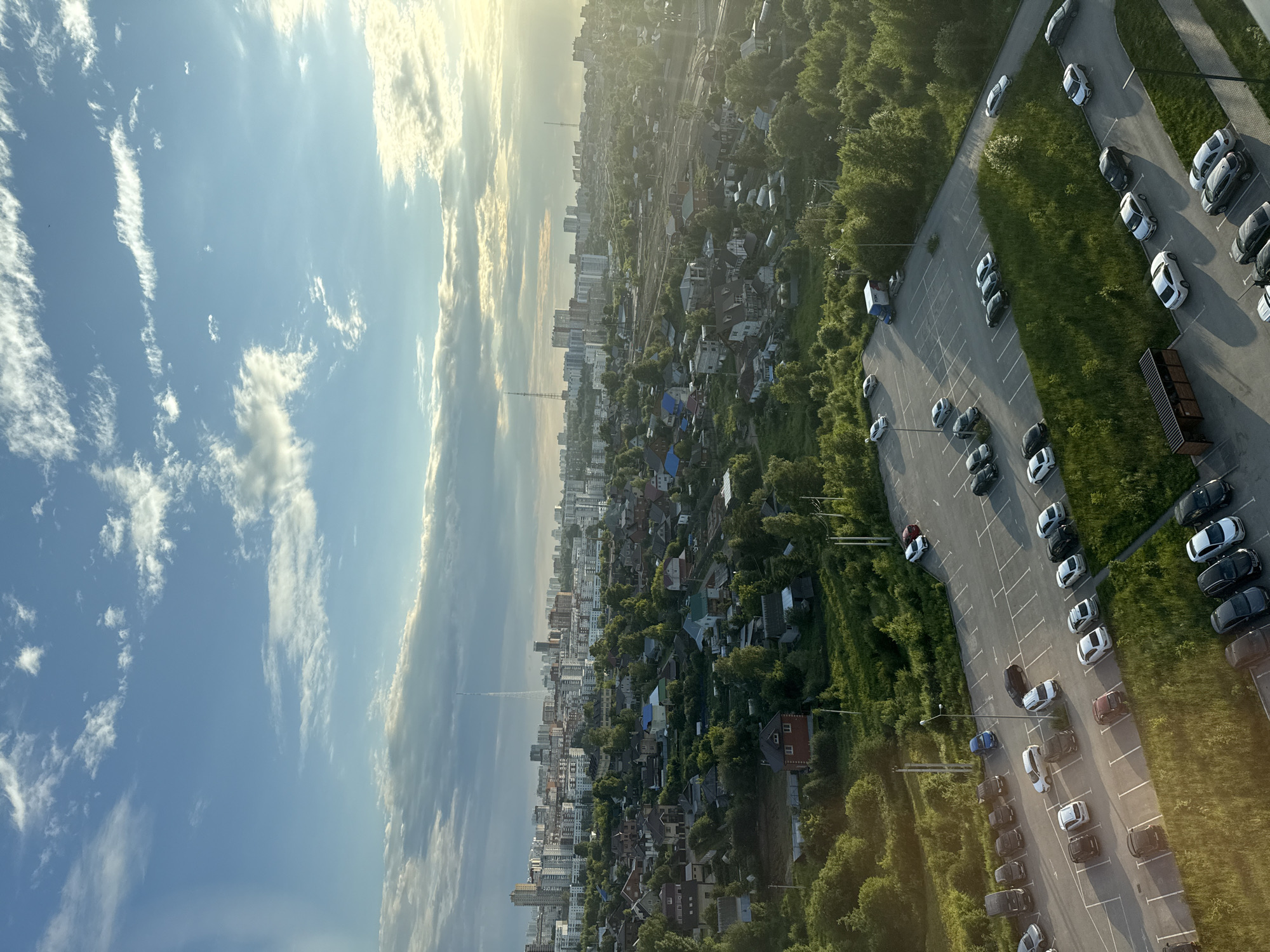 This is a high-angle aerial shot of a residential and urban area, likely taken during the late afternoon or early evening, with the sun setting to the right, casting long shadows and creating a warm glow. The foreground features a large, paved parking lot filled with numerous cars of various colors and models, some parked neatly in designated spots, others more haphazardly. Adjacent to the parking lot is a grassy area with some trees.
Beyond the parking lot, the scene transitions to a sprawling residential neighborhood characterized by numerous houses with red-tiled roofs and green trees. Further in the distance, a dense cityscape emerges, dominated by numerous multi-story apartment buildings and some taller structures that appear to be communication towers or residential high-rises. The overall impression is one of a densely populated urban environment interspersed with green spaces and a lower-density suburban or fringe area.
Based on the visual cues, particularly the architectural style of the buildings, the presence of communication towers with distinctive lattice structures, and the overall urban layout, this scene is likely located in the city of Perm, Russia. Specifically, the view appears to encompass a transition zone between a suburban or dacha settlement and a more developed urban core. The presence of a large parking lot serving a residential or commercial area suggests a connection to the wider city infrastructure.