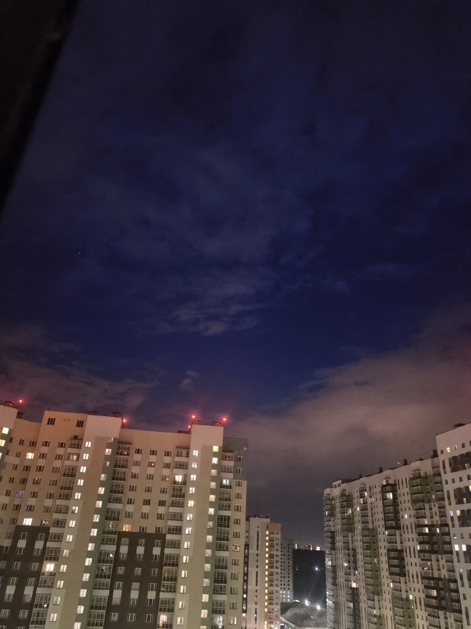 The image depicts a nighttime cityscape with multiple residential buildings illuminated by interior lights. The sky above is a deep blue with scattered clouds, suggesting it is dusk or night. Red lights are visible on top of some of the buildings, possibly indicating warning lights for aircraft. The buildings themselves are tall and modern, with numerous windows showing various levels of illumination, hinting at occupied apartments. The perspective is from a higher vantage point, looking down at the tops of some buildings and across at others. There are no people or specific objects clearly visible outside of the buildings and the sky. The scene evokes a sense of urban living at night, with the quiet hum of a city after dark. The weather appears to be clear enough for the sky to be visible, though the clouds suggest some atmospheric activity. The overall mood is calm and somewhat detached, typical of a nocturnal urban view. No text is discernible in the image.