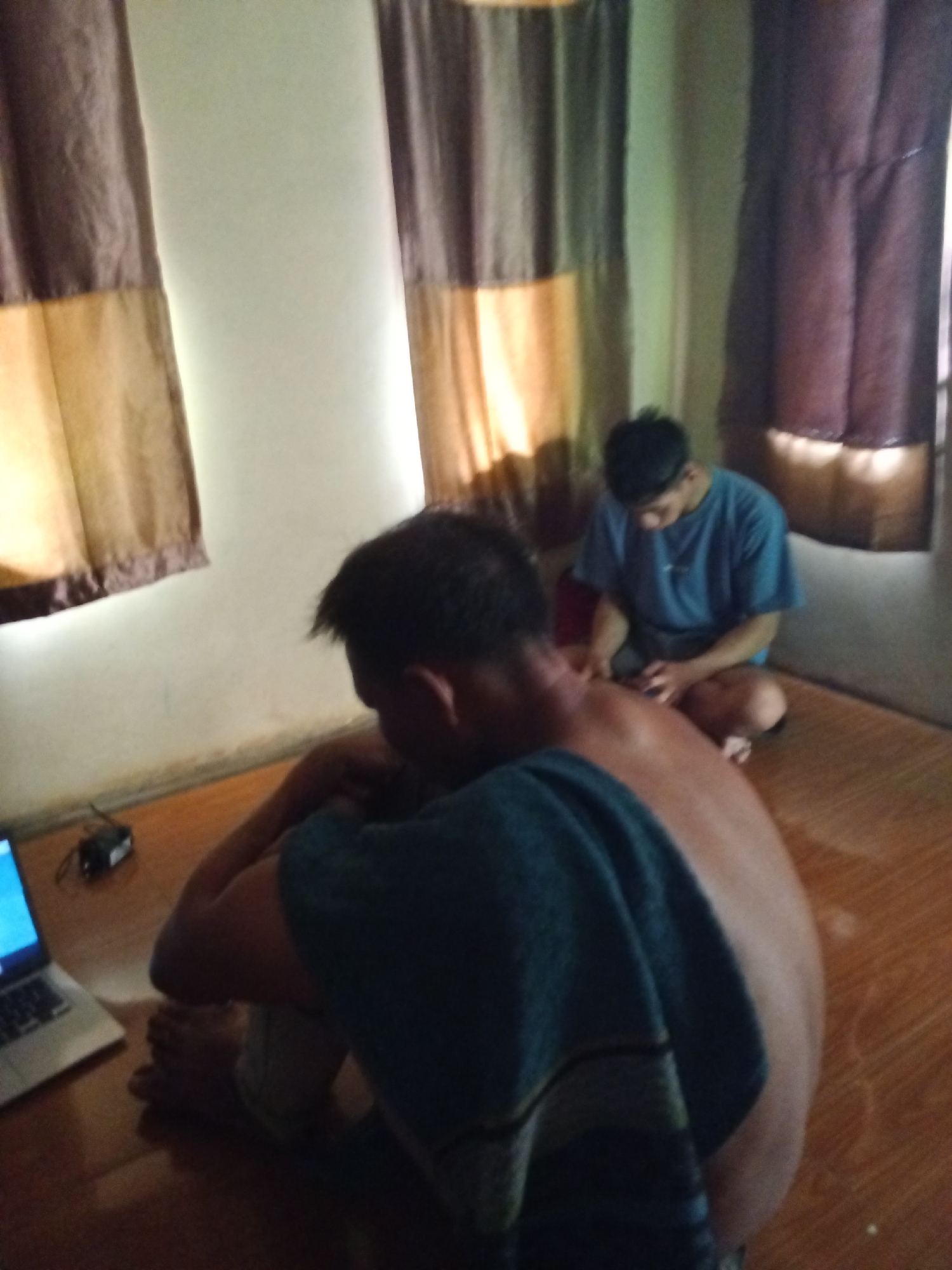 The image depicts two young men in what appears to be a sparsely furnished room. One man is in the foreground, seen from the back, with his head bowed and wearing a dark blue t-shirt. He is sitting on the floor, with a laptop open in front of him on a wooden surface. A towel or blanket is draped over his shoulders and upper back. The other man is in the background, sitting cross-legged on the floor and looking down at his hands, appearing to be engaged with a small object or device. He is wearing a light blue t-shirt and shorts. The room has light-colored walls and wooden floorboards. There are two large windows with curtains that appear to be made of different colored fabric panels, suggesting they are decorative or serve to block light. The lighting in the room is dim, with some light filtering through the curtains, creating a warm glow. The overall impression is one of casual indoor activity. The presence of a laptop and the general setting do not provide specific clues to identify a precise location within Subang, Indonesia, but it could be a residential room or a small business establishment.