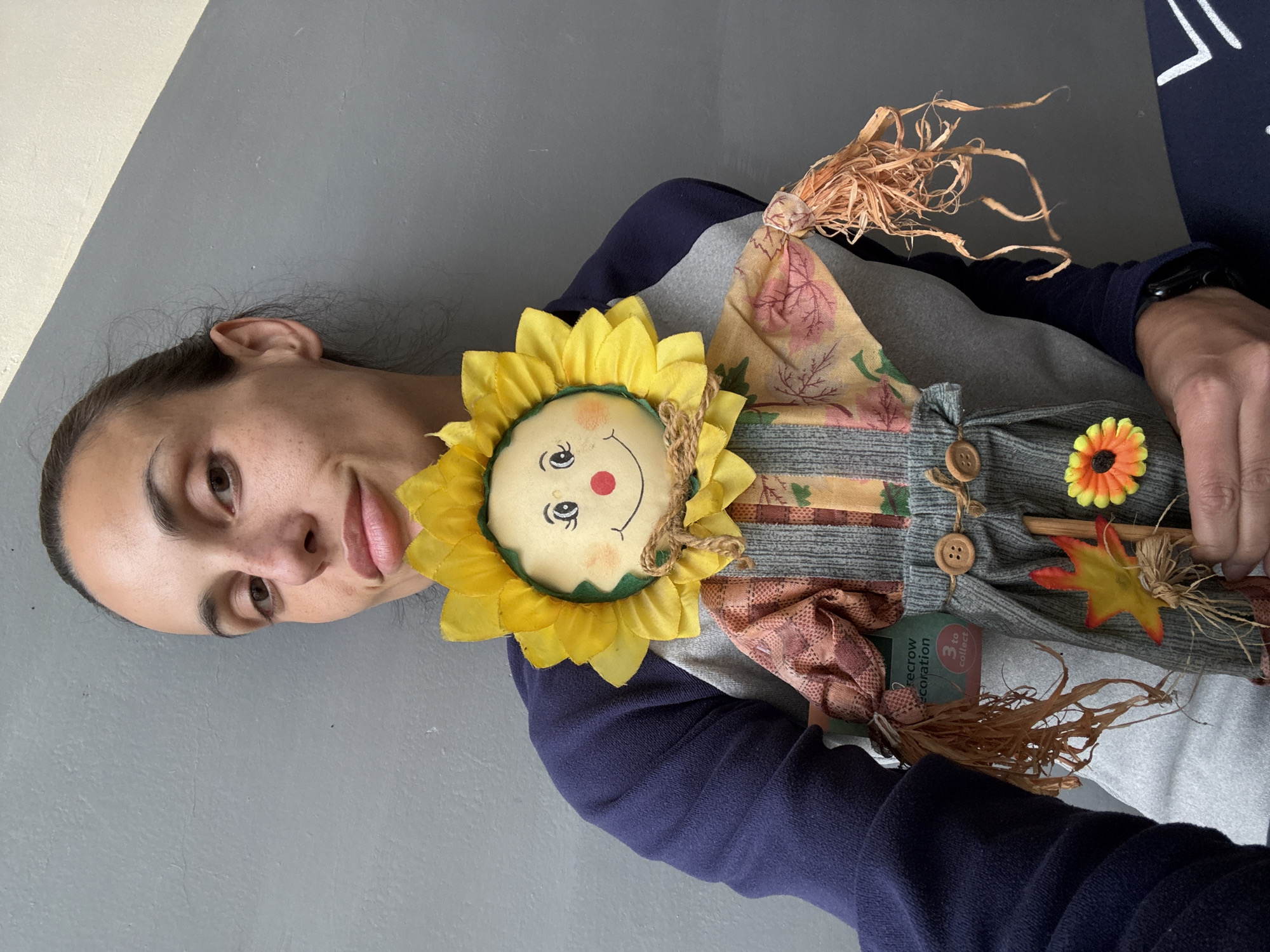 A person is depicted holding a decorative scarecrow against a two-toned wall, split diagonally between a lighter off-white and a darker gray. The image is oriented sideways, with the person's head positioned on the left. The individual has dark hair pulled back, brown eyes, and a neutral to slightly smiling expression. They are wearing a long-sleeved garment, possibly a sweatshirt, with dark blue sleeves and a lighter grey body. A black smartwatch is visible on their left wrist.
The scarecrow decoration, held prominently by the person, features a cheerful sunflower head made of yellow fabric petals surrounding a beige center with embroidered smiling facial features (black eyes, red nose, red smile). Its body is composed of various fabric patches, including patterns of autumn leaves and stripes, and is embellished with small wooden buttons. Tufts of straw, resembling hair or limbs, extend from the decoration. Additional adornments include a small orange and yellow fabric flower and a red and yellow artificial maple leaf. A visible tag on the decoration reads "SCARECROW DECORATION" and "3 to collect." The setting appears to be indoors in Pleven, Bulgaria, under even lighting.
