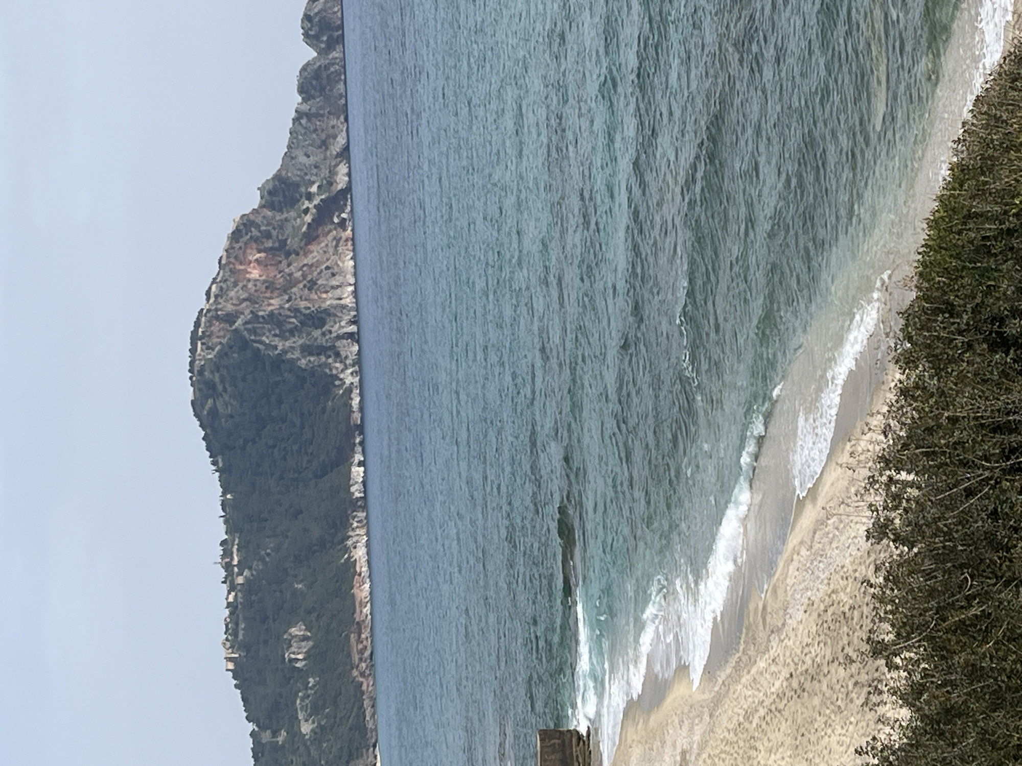 The image depicts a coastal scene in Alanya, Turkey. The horizon is nearly vertical and in the middle of the frame. On the left, a mountain rises, covered in vegetation, with some rocky, reddish-brown areas near the peak. Scattered buildings are visible on the mountain's slope. The majority of the frame is occupied by the sea, displaying varying shades of blue. Waves are breaking on a sandy beach in the bottom right corner, with white foam creating a border between the water and the shore. Dense green foliage is located along the right edge of the frame, partially obscuring the beach. In the bottom center of the frame are some rocky formations in the water. The sky is partially visible in the upper left corner. The lighting appears soft, suggesting either a cloudy day or shooting near dawn or dusk.