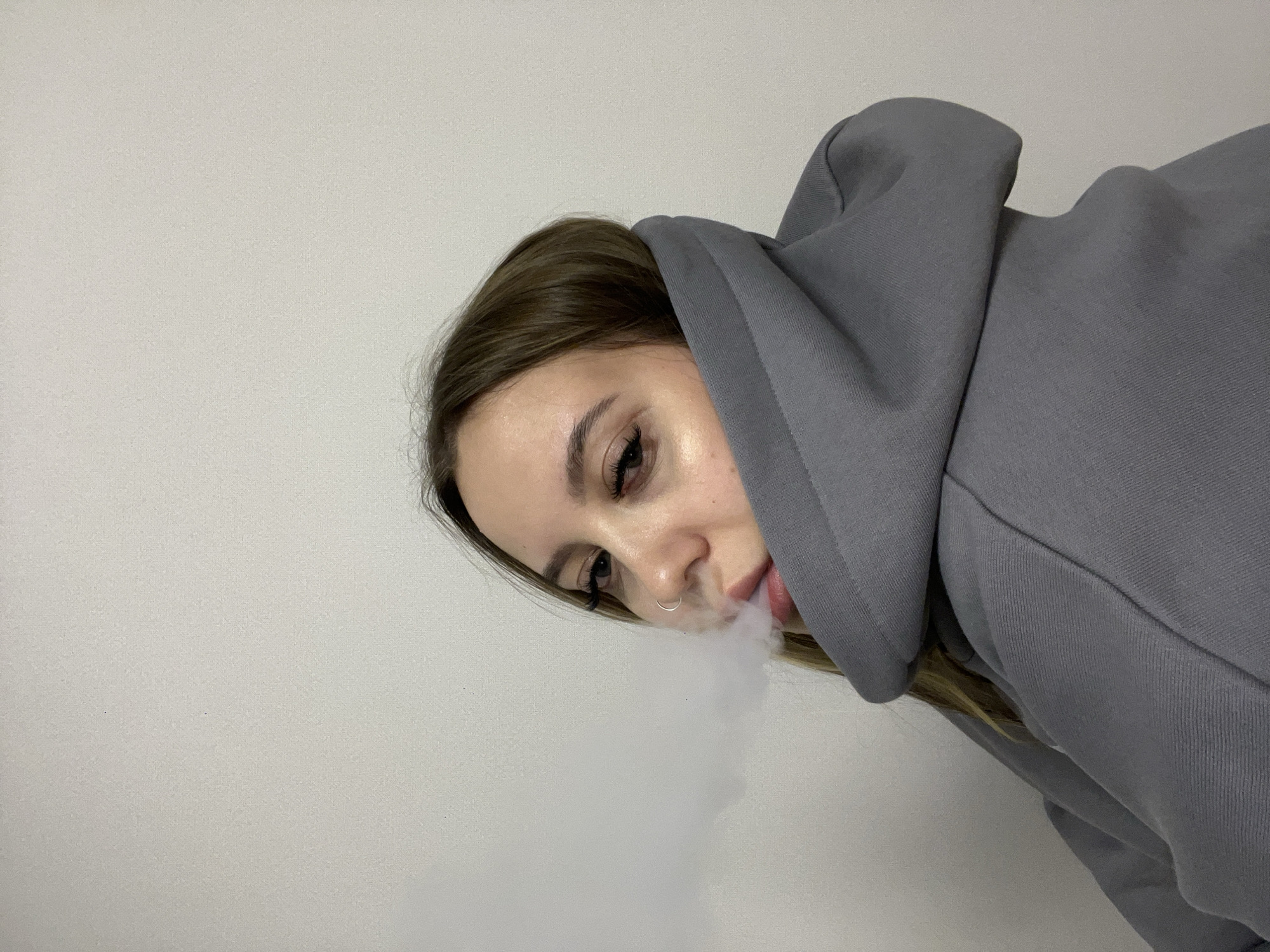 A medium close-up, angled photograph features a young woman from the chest up, positioned against a plain, light gray background, likely indoors in Krasnodar, Russia. She has long brown hair, partially visible, with the rest covered by the hood of a simple gray hooded sweatshirt. The hood is up, framing the right side of her face. The woman is actively exhaling a prominent cloud of white vapor from her mouth, indicating she is vaping or smoking. Notable details include her defined eyelashes, possibly extensions, and a small silver hoop nose ring. Her gaze is directed downwards and to the left. The lighting is even and appears to be artificial or diffuse natural light. There is no visible text or specific time of day indicated in the image. The overall scene is casual, with the focus on the woman and the vapor she is exhaling.