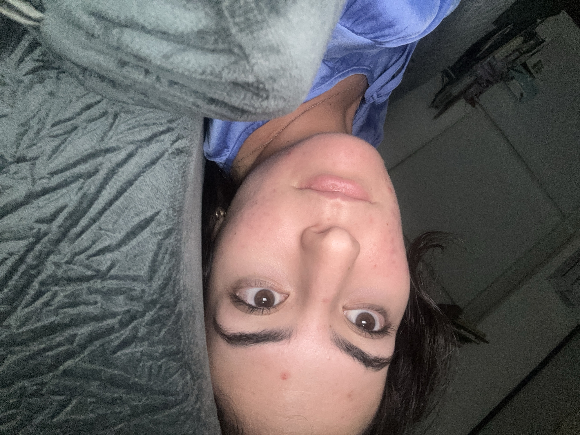 A close-up, rotated view of a person lying down. The person has fair skin, brown eyes, and dark eyebrows. They are wearing a blue shirt and a thin necklace. Part of a gray blanket or sheet is visible near their left side. The background is dark, suggesting a dimly lit room. The location is not discernible from the image.