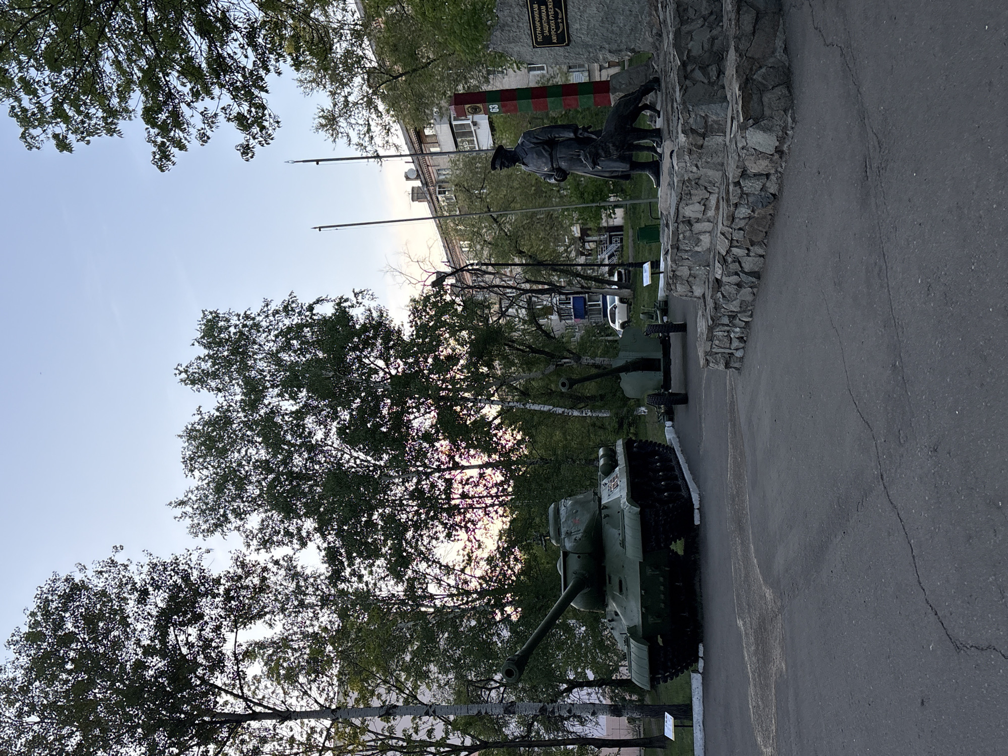 The image shows a war memorial in Birobidzhan, Russia. There is a green tank and cannon. A statue of a soldier and dog can be seen behind a border marker. The memorial appears to be outside along a road with trees in the background.