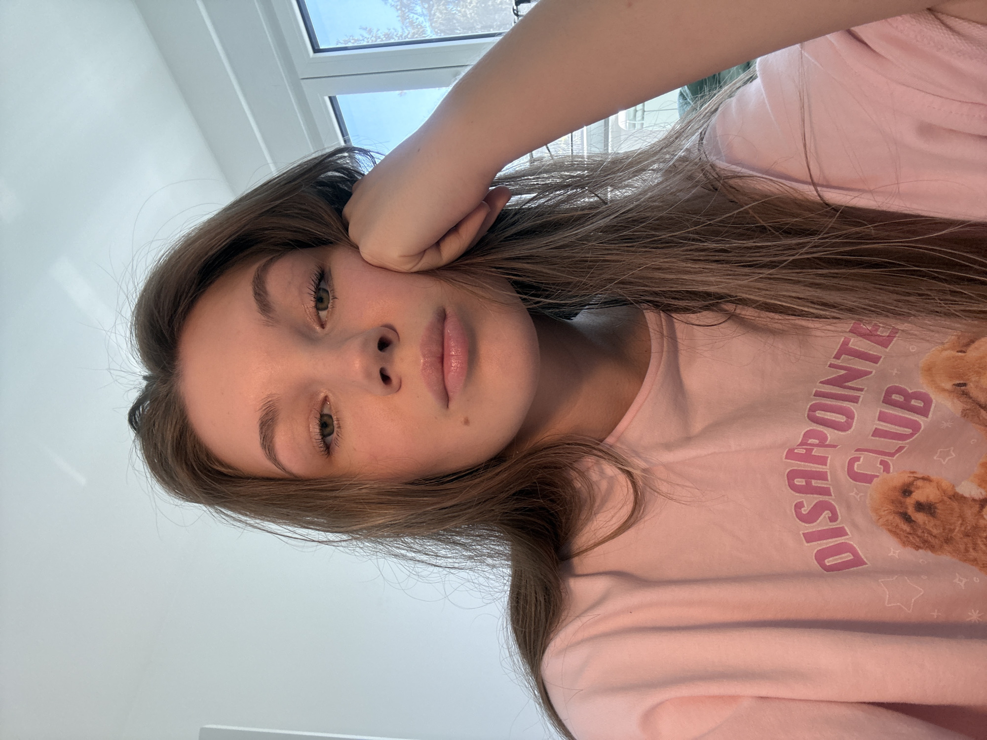 A young woman is indoors. She is wearing a pink shirt with the words "DISAPPOINTED CLUB" printed on it. The woman has long, brown hair and is resting her head on her hand. The background is a white wall with a window. The location is not discernible from the image.