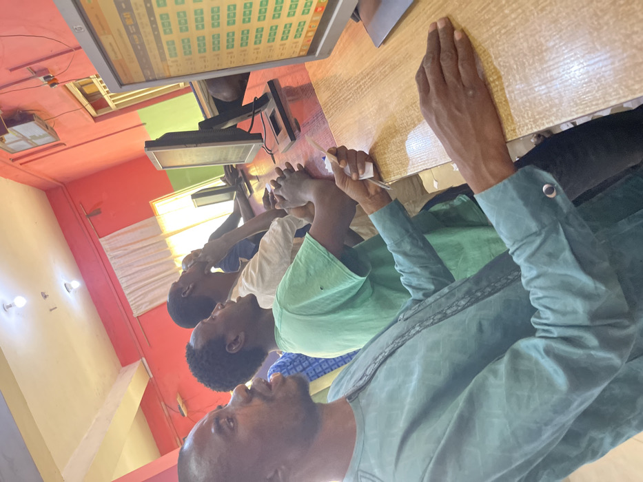 This image depicts an interior scene within a computer facility or internet cafe in Chikaji, Nigeria. Four adult males are visible, positioned along a continuous wooden counter. The room features reddish-orange walls, a light-colored ceiling with two illuminated bare light bulbs, and white-curtained windows in the background. The closest male, wearing a light teal patterned long-sleeved garment, holds a small white paper slip in his right hand while resting both hands on the wooden counter, his gaze directed forward towards a computer monitor. This monitor displays a grid of green squares with a "00:41" timer. Behind him, a male in a green t-shirt has his hands clasped on the counter, also looking forward. Further down the counter, a third male in a light-colored shirt is visible, with one hand touching his chin as he looks towards a screen. A fourth individual is partially visible. Multiple black-framed computer monitors are present on the counter.