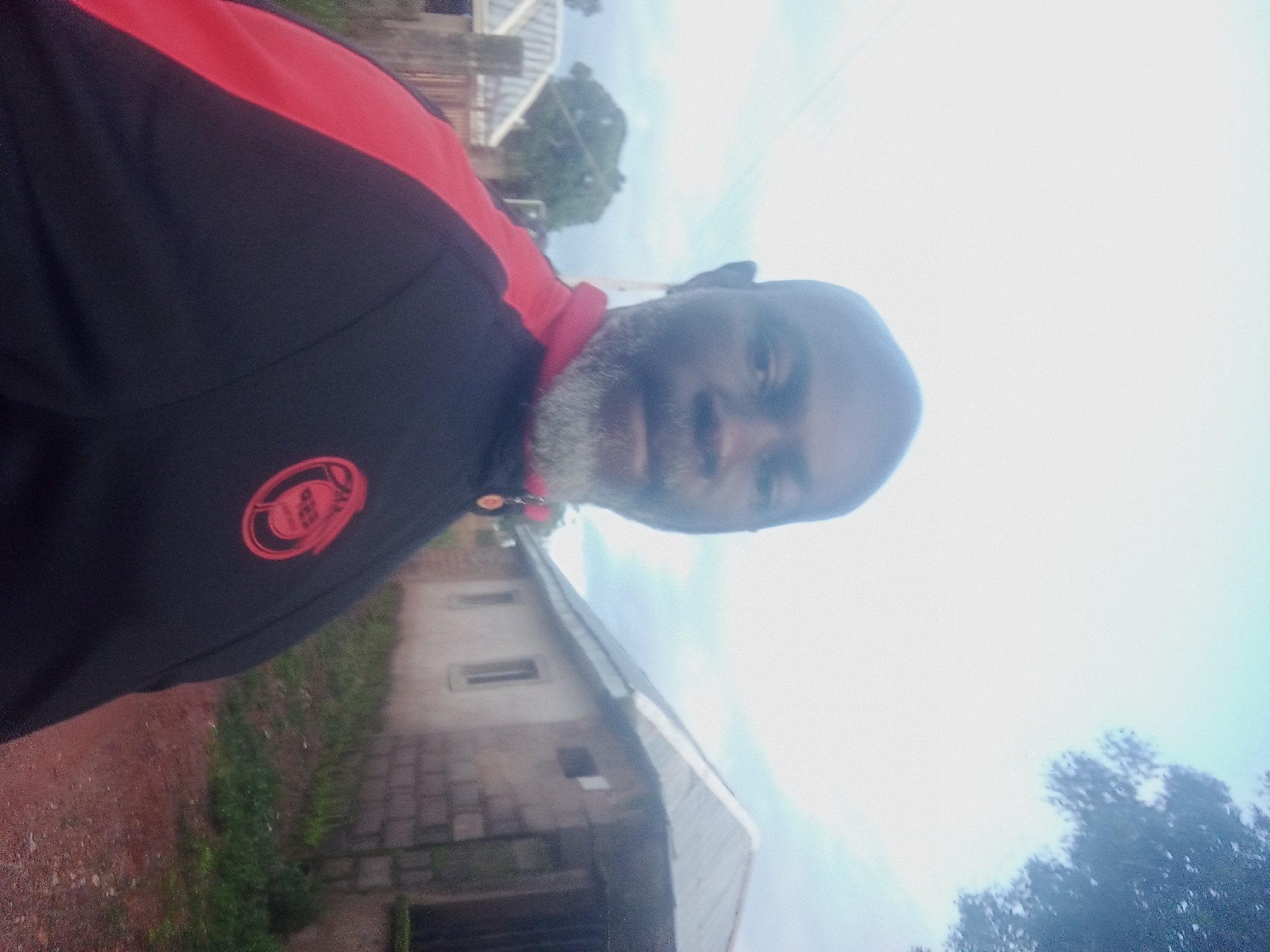 A middle-aged man with a salt-and-pepper beard stands in front of a house with a corrugated metal roof in Rafin Bauna, Nigeria. He is wearing a black jacket with red accents. The house is constructed from concrete blocks and has a rectangular window. The ground is a dirt path. The sky is overcast, with some tree branches visible in the upper right corner.