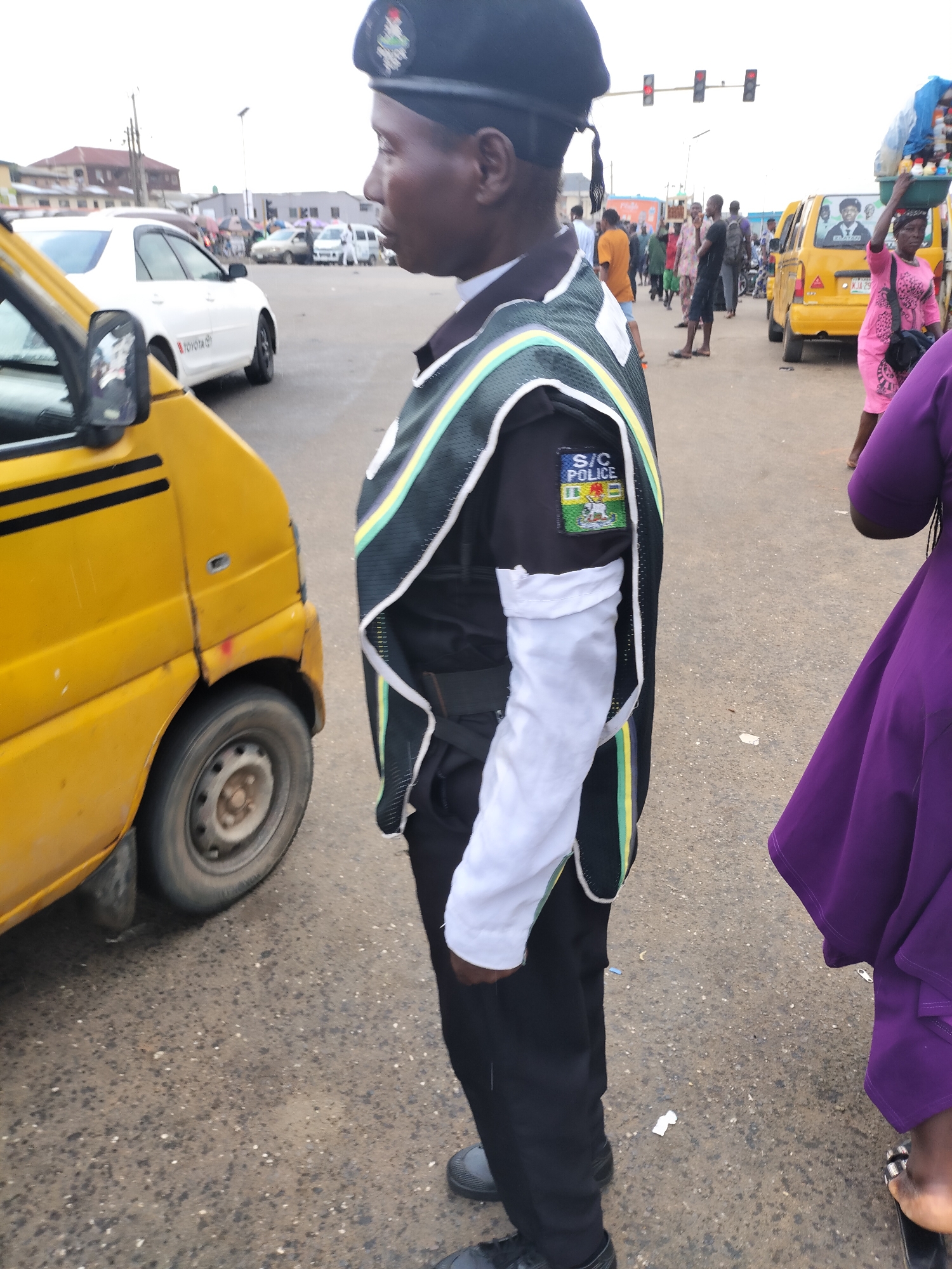 The image depicts a traffic police officer in Ikorodu, Nigeria, standing at a busy intersection. He wears a black uniform, a black beret, and a vest with green, yellow, and white stripes. The patch on his left sleeve reads "S/C Police." A yellow taxi is parked to his left, and people cross the street in the background. A woman in a purple dress stands to his right. The scene shows general street activity with other vehicles and pedestrians in the background under overcast lighting. Traffic lights are visible overhead.