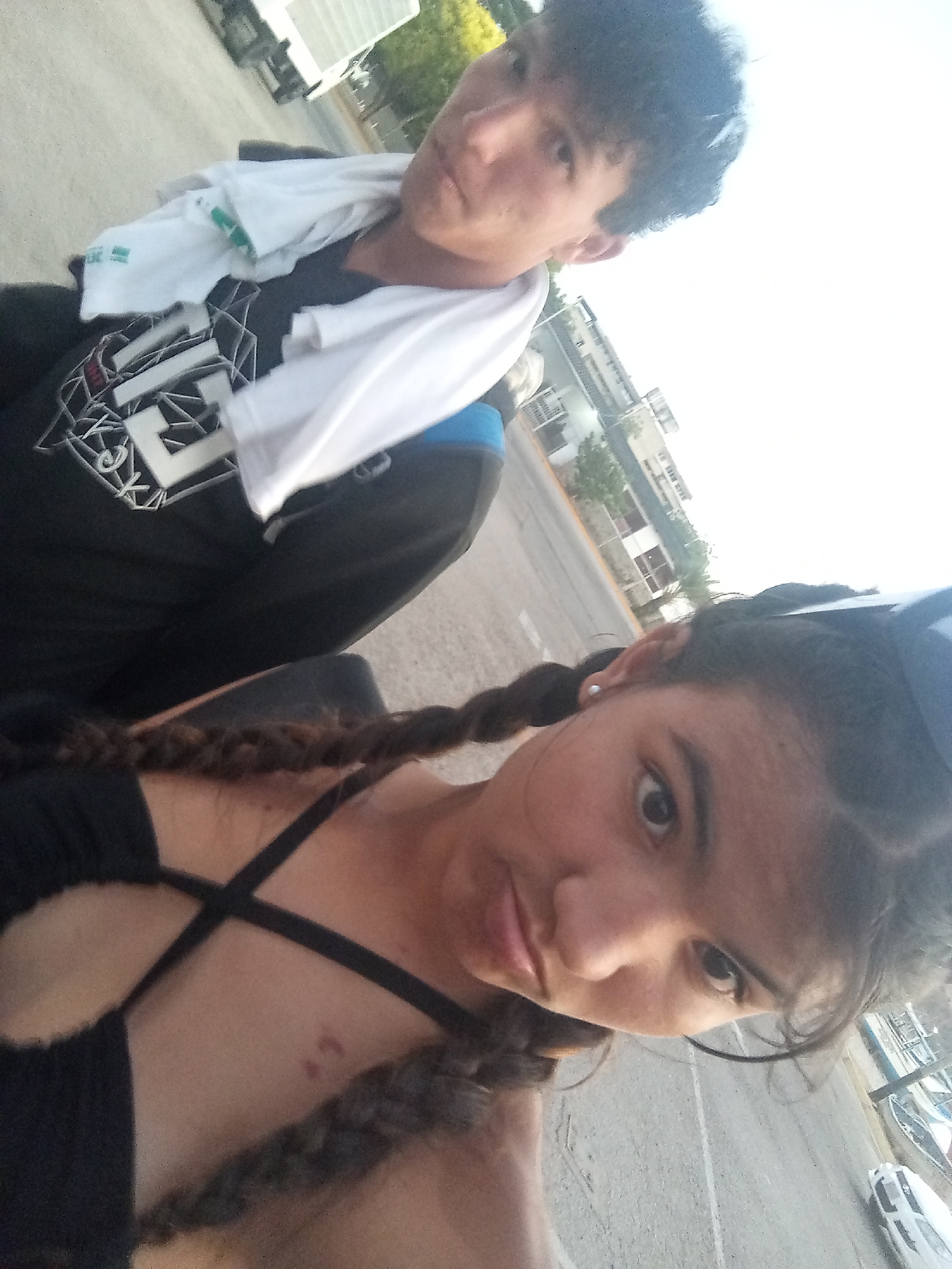 A medium, angled-upward shot shows two individuals outdoors. The person in the foreground is a young female with dark hair styled in braids, wearing a black top with crossed straps. The individual behind her is a young male with short, dark curly hair. He is wearing a black shirt with a white design and a white towel around his neck. The setting appears to be an outdoor space in Juan Griego, Venezuela. In the background, a road, buildings, and some vehicles are visible. The overall impression is of a casual, outdoor setting with two individuals.