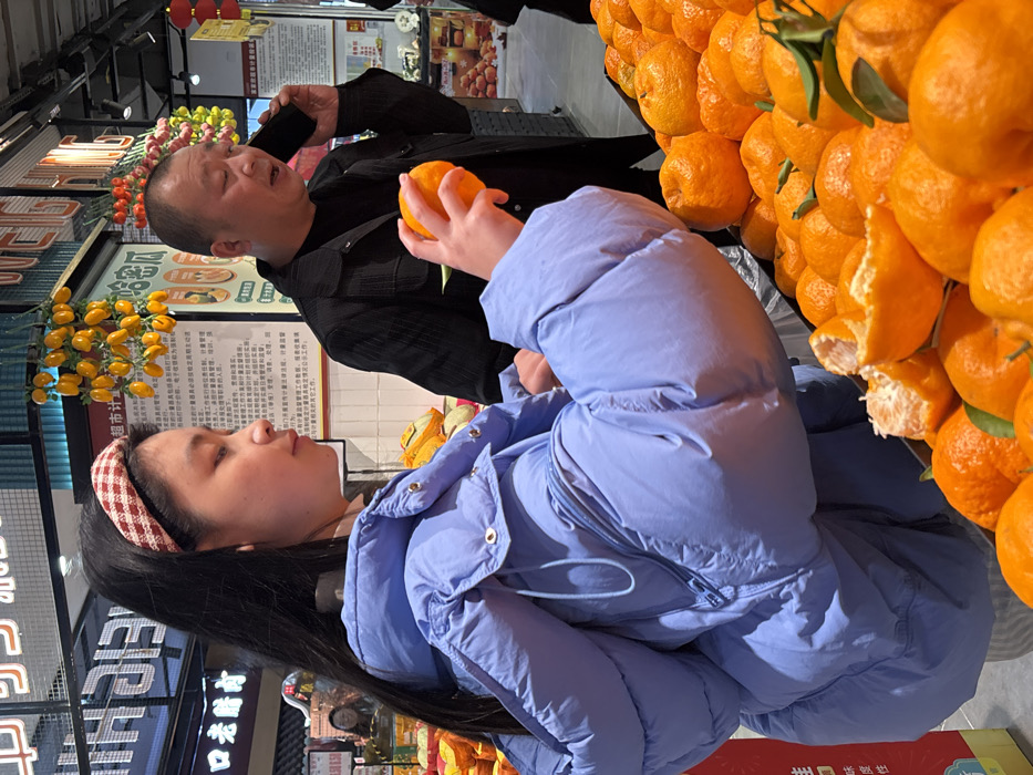 The scene is set indoors within a supermarket or grocery store produce section in Jiangjin, China. A young female, positioned in the foreground and facing right, has long dark hair and wears a red and white checkered headband. She is dressed in a light blue puffy jacket and holds an orange in her right hand, which she is actively peeling, directing her gaze downwards toward a large, elevated pile of oranges and tangerines. One orange within this pile is already partially peeled, revealing its segments. Directly behind her, in the mid-ground, a middle-aged male with short dark hair, wearing a dark jacket, holds a black smartphone to his left ear and appears to be speaking into it. His mouth is open, and his attention is directed away from the female. The background features various supermarket elements including vertical and horizontal signage with Chinese characters, overhead lighting, and other produce displays, notably decorative yellow gourd-like items hanging from the ceiling and red lantern-like ornaments. A faint reflection of another individual is visible in a distant, reflective surface on the far left.
