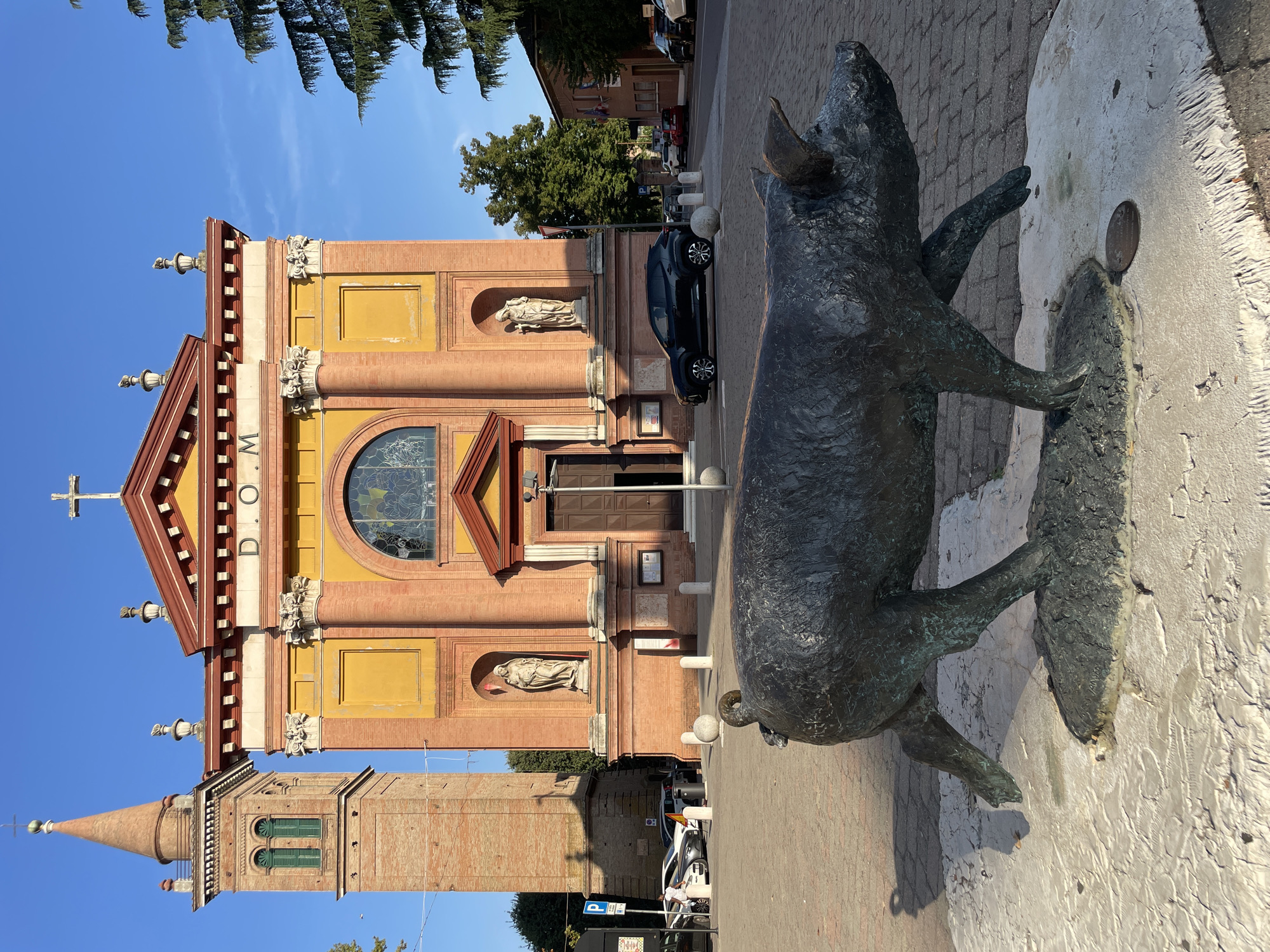 The image captures the exterior of the Church of the Immaculate Conception in Castelnuovo Rangone, Italy, with a bronze statue of a pig prominently placed in the foreground. The church features a yellow facade with red brick detailing and columns, topped with a red roof and a cross. To the left of the church is a tall, slender red brick bell tower with green shutters on its windows. The statue of the pig is dark bronze and appears to be in motion, positioned on a light-colored, textured base. The scene is set on a sunny day, with a clear blue sky. In the background, there are parked cars, streetlights, trees, and other buildings, suggesting an urban or town square environment. The cobblestone pavement is visible in front of the church and around the statue.